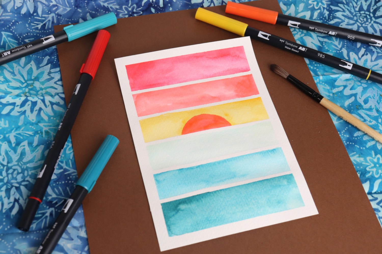 Image contains a postcard sized paper colored with six horizontal stripes to form a sunset. A variety of colored markers and a paintbrush lay around it on a blue batik fabric background.