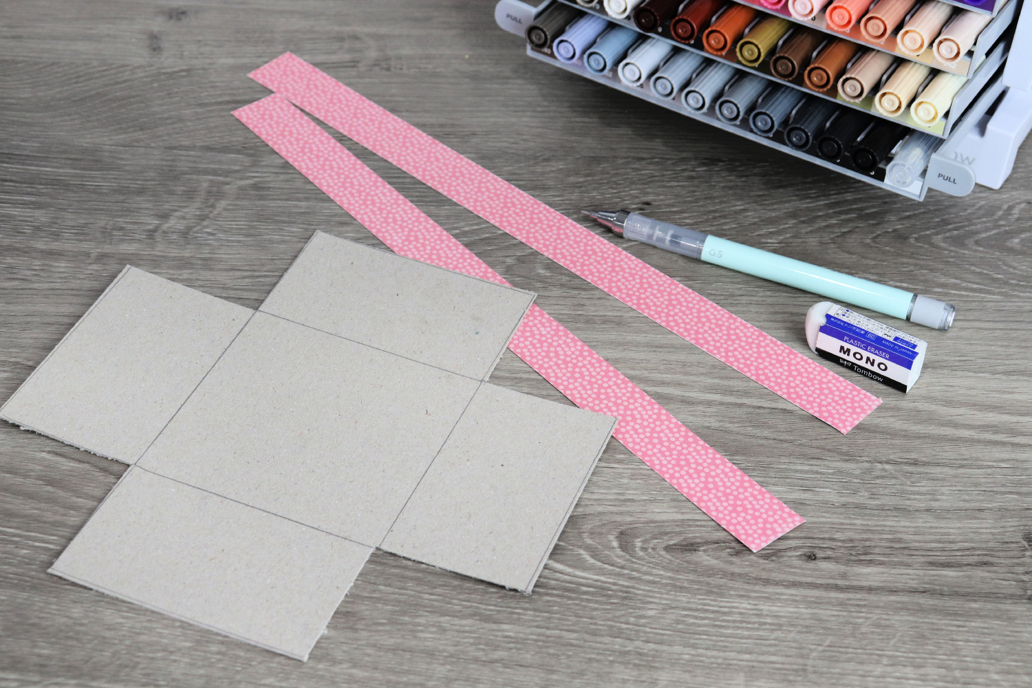 Image contains a piece of cut chipboard and two scrapbook paper strips on a wooden desk. A mechanical pencil, an eraser, and a marker organizer sit nearby.