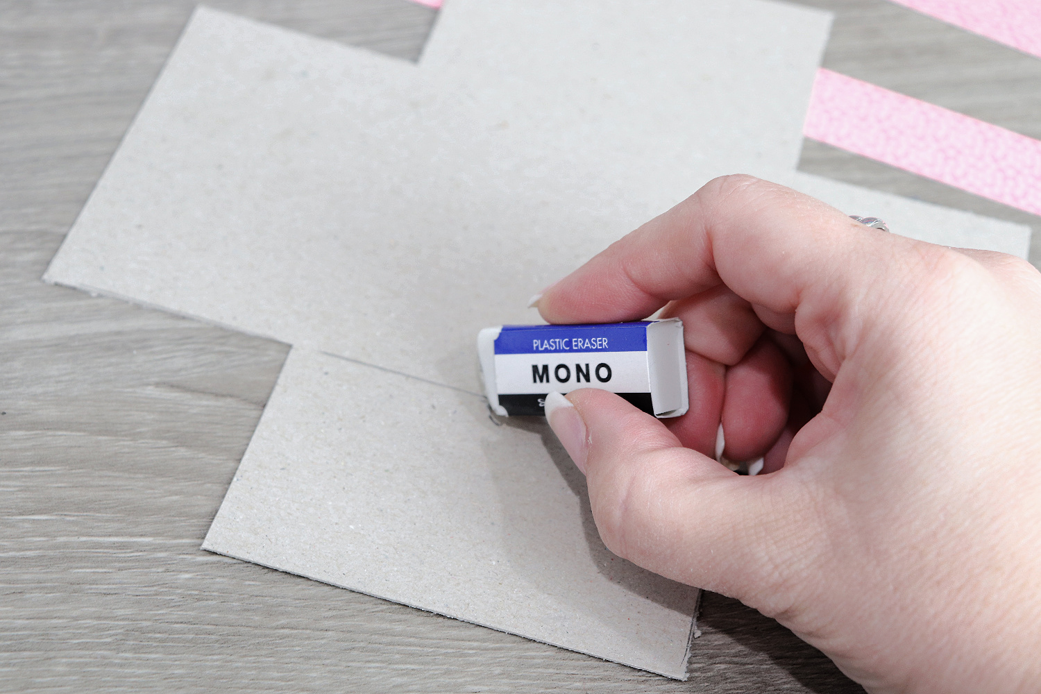 Image contains Amy’s hand holding an eraser over a cut piece of chipboard laying on a wooden desk.