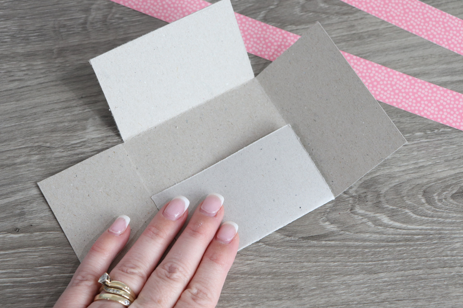 Image contains Amy’s hand folding a piece of chipboard on a wooden desk. Two strips of pink patterned scrapbook paper sit nearby.