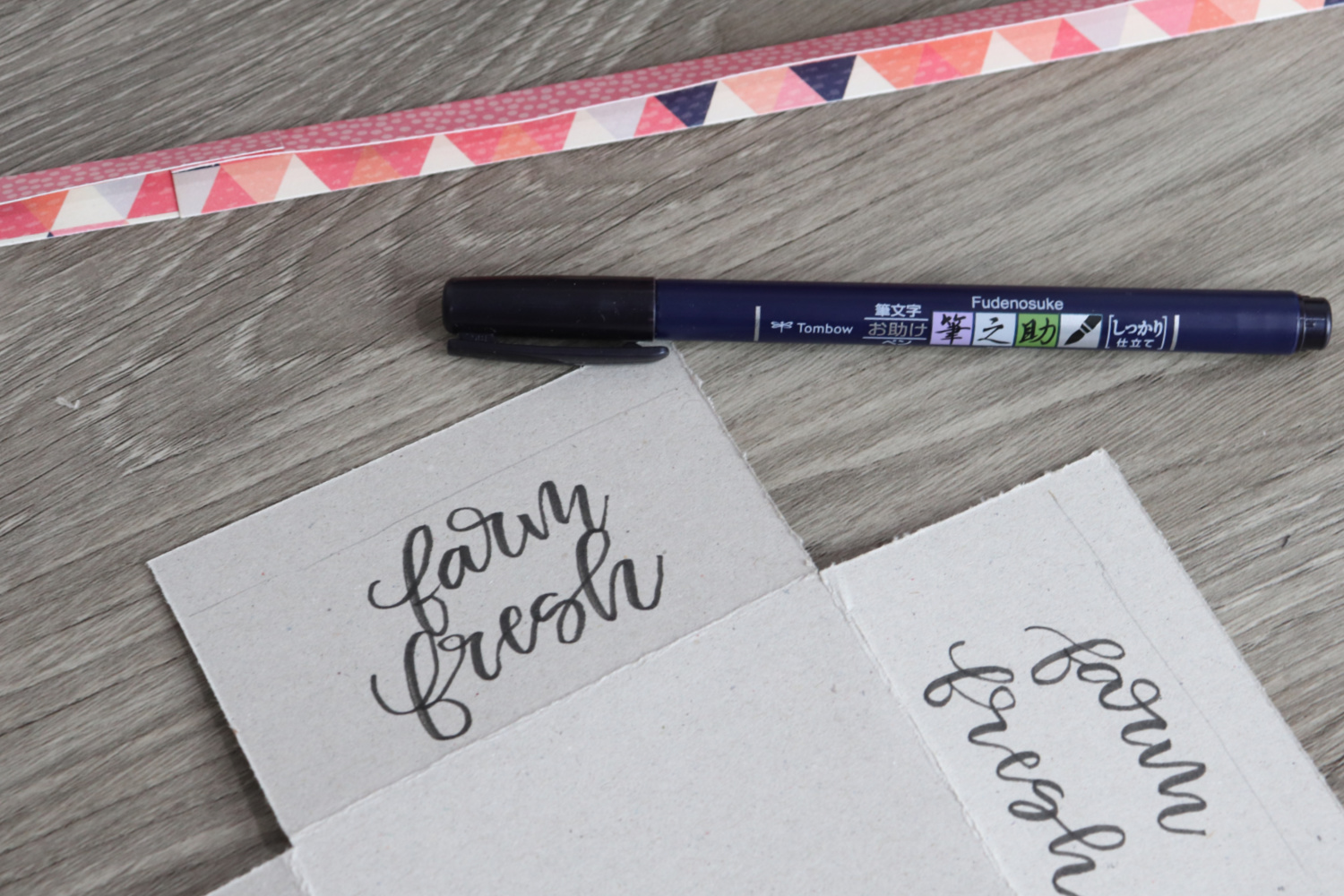 Image contains a piece of chipboard with “farm fresh” written on it. A Fudenosuke pen and a folded piece of scrapbook paper sit nearby on a wooden table.