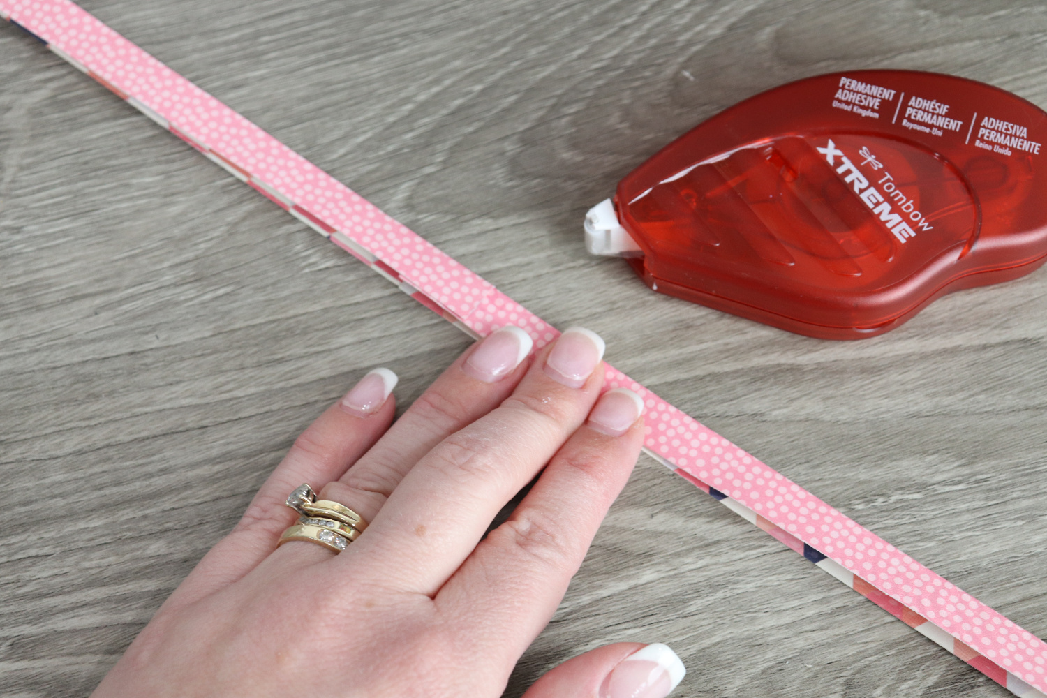 Image contains Amy’s hand folding a piece of pink patterned scrapbook paper. An Xtreme Adhesive Runner sits nearby on a wooden table.