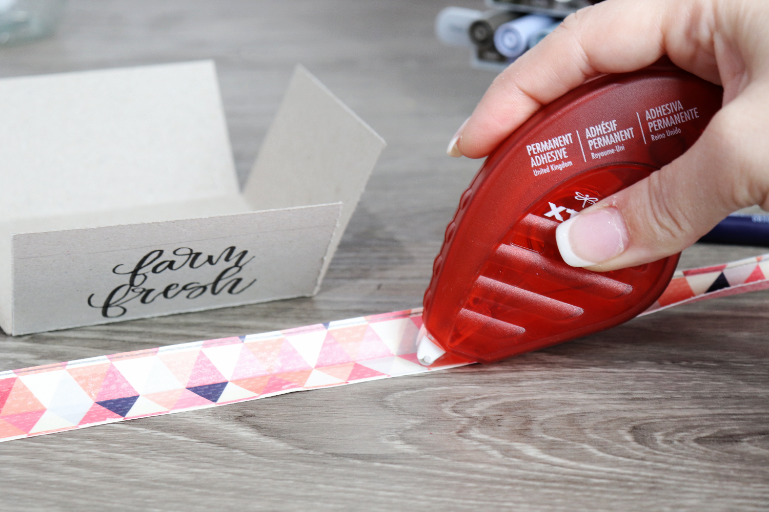 Image contains Amy’s hand applying an adhesive tape runner to a piece of scrapbook paper. A chipboard box sits in the background on a wooden table.
