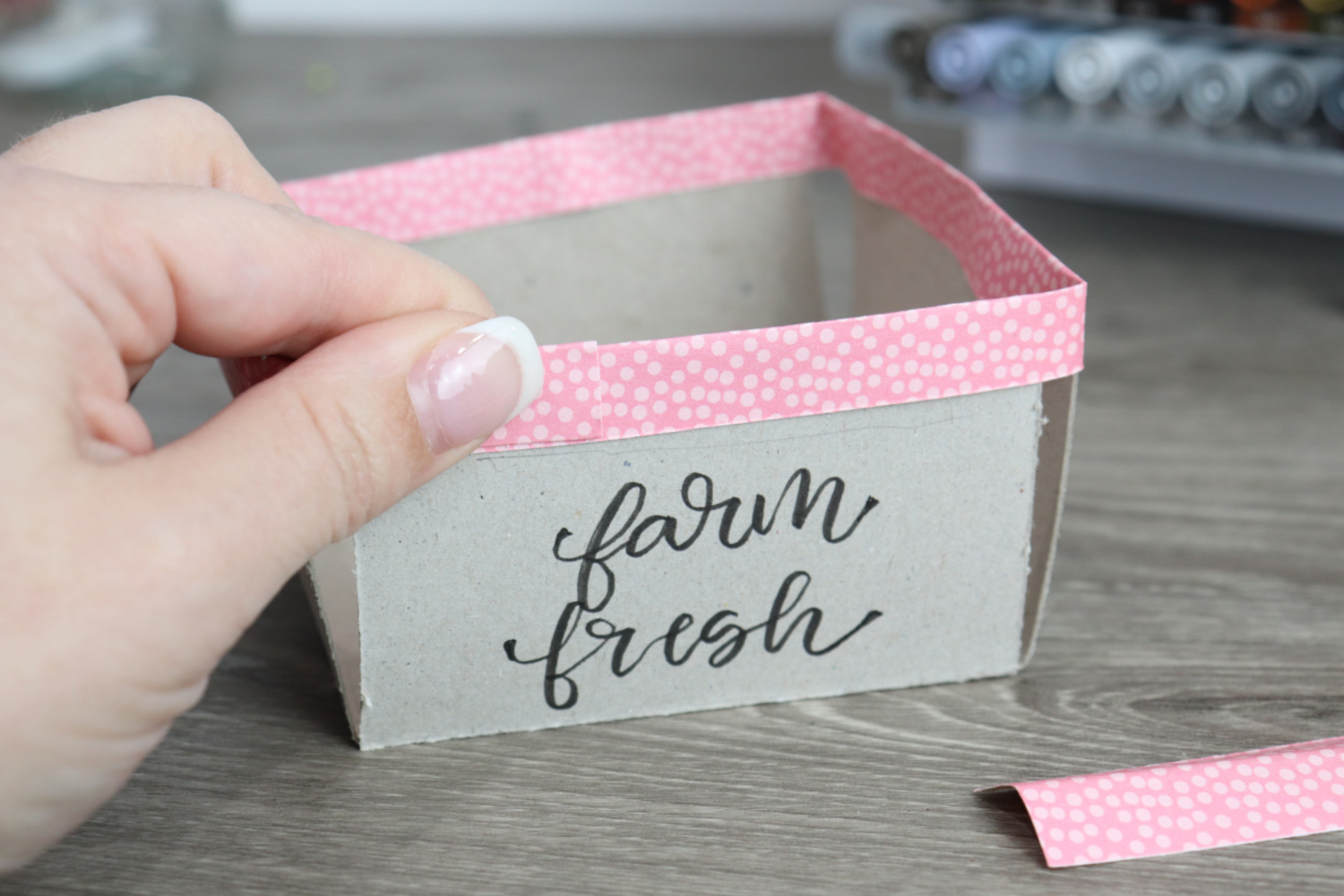 Image contains Amy’s hand applying a pink scrapbook paper strip to the top of a chipboard box labeled “farm fresh.” 