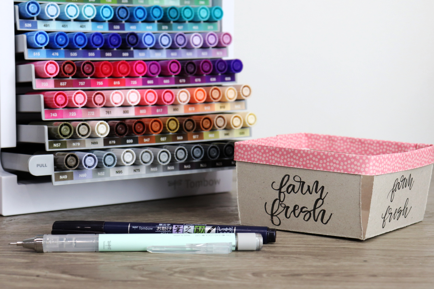 Image contains a chipboard berry basket sitting on a wooden desk next to a marker organizer. A brush pen and a mechanical pencil sit nearby.