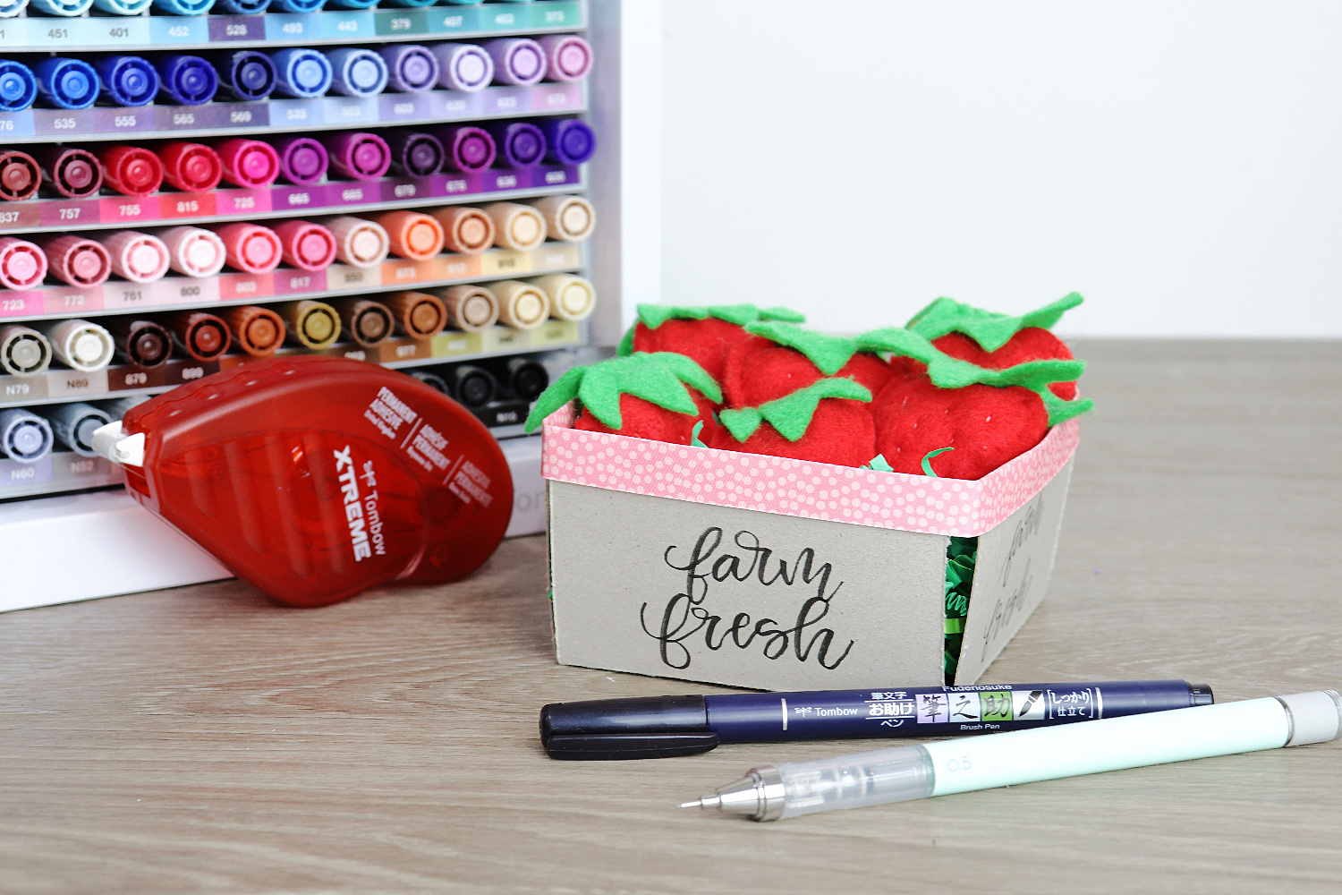 Image contains a chipboard berry basket filled with small felt strawberries. An organizer filled with colored markers, an Xtreme Adhesive Tape Runner, a Fudenosuke Pen, and a mechanical pencil sit nearby on a wooden table.
