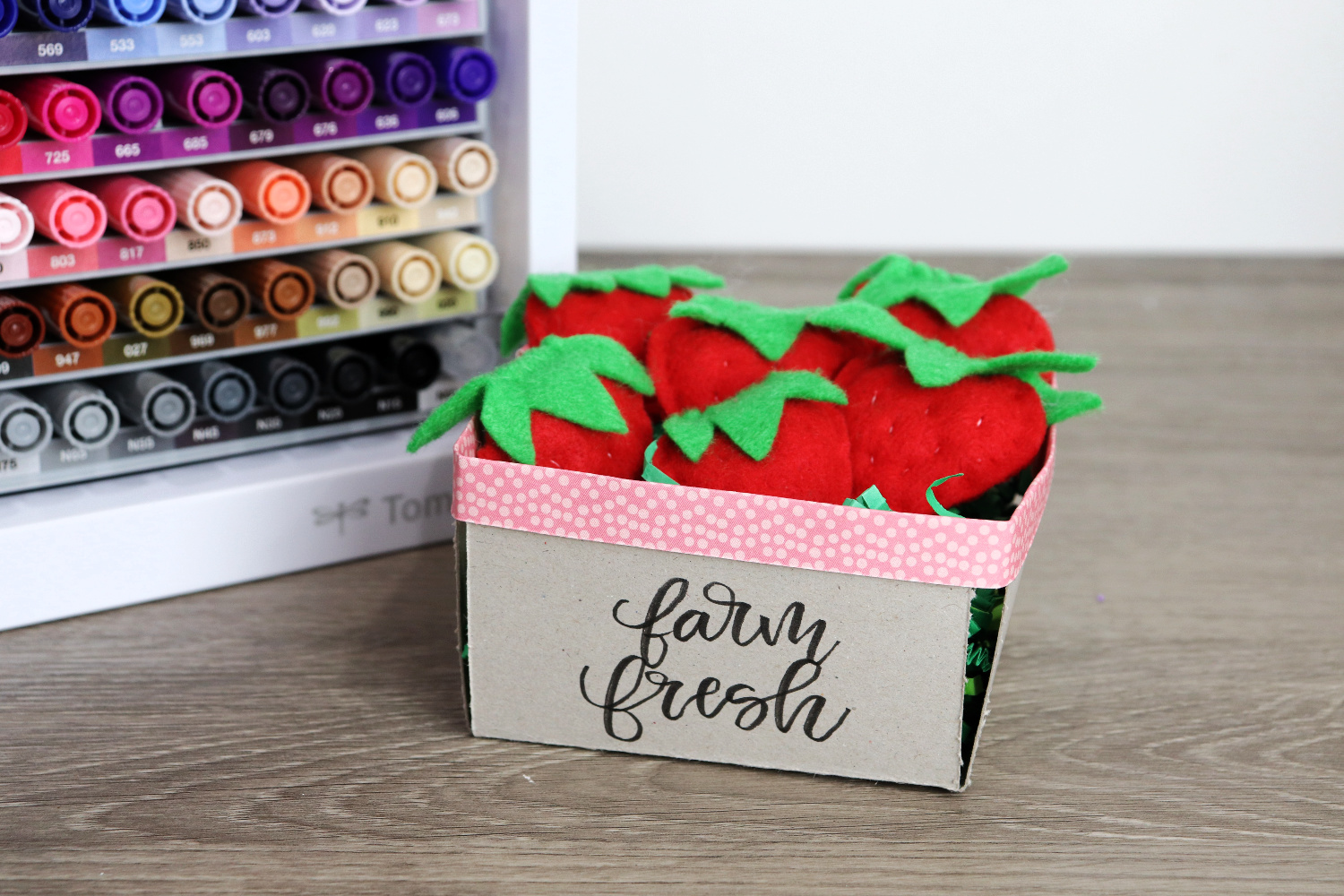 Image contains a chipboard berry basket filled with small red felt strawberries sitting on a wooden desk. A marker organizer sits nearby. 