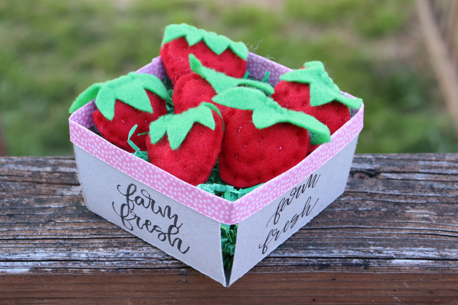 Image contains a chipboard berry basket filled with red felt strawberries sitting on a wooden railing.