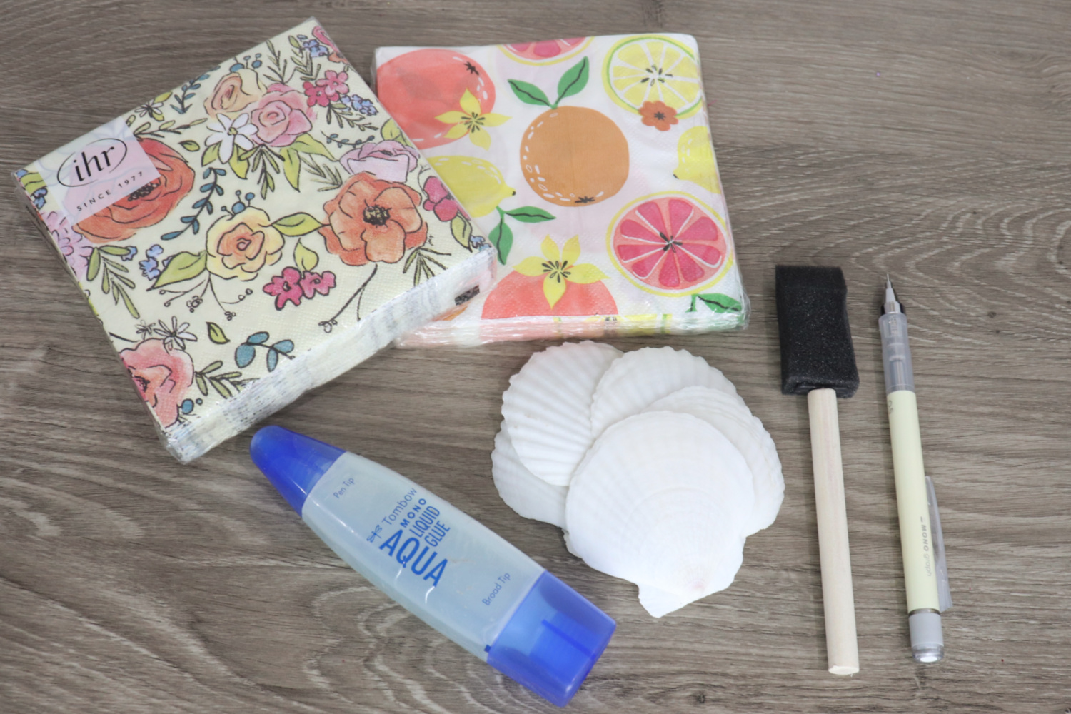 Image contains two packs of patterned napkins, a tube of Tombow MONO Aqua Liquid Glue, five large white scallop shells, a sponge brush, and a mechanical pencil on a wooden desktop.