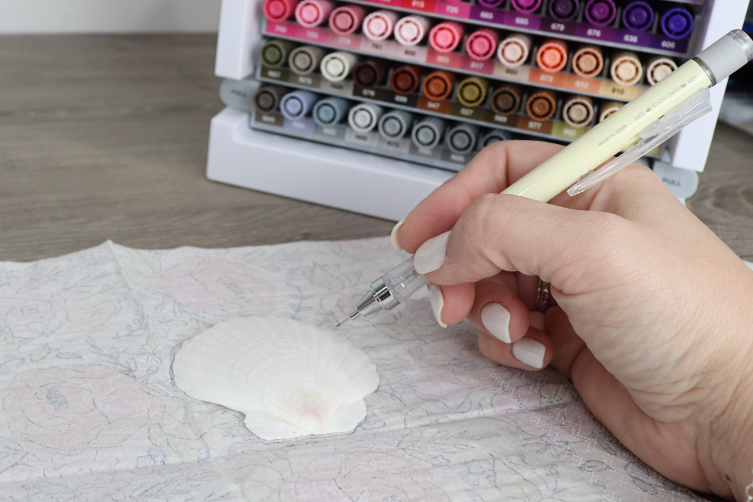 Image contains Amy’s hand holding a MONO Graph mechanical pencil and tracing a large white scallop shell onto the back of a patterned napkin. A marker organizer sits nearby on a wood desktop.