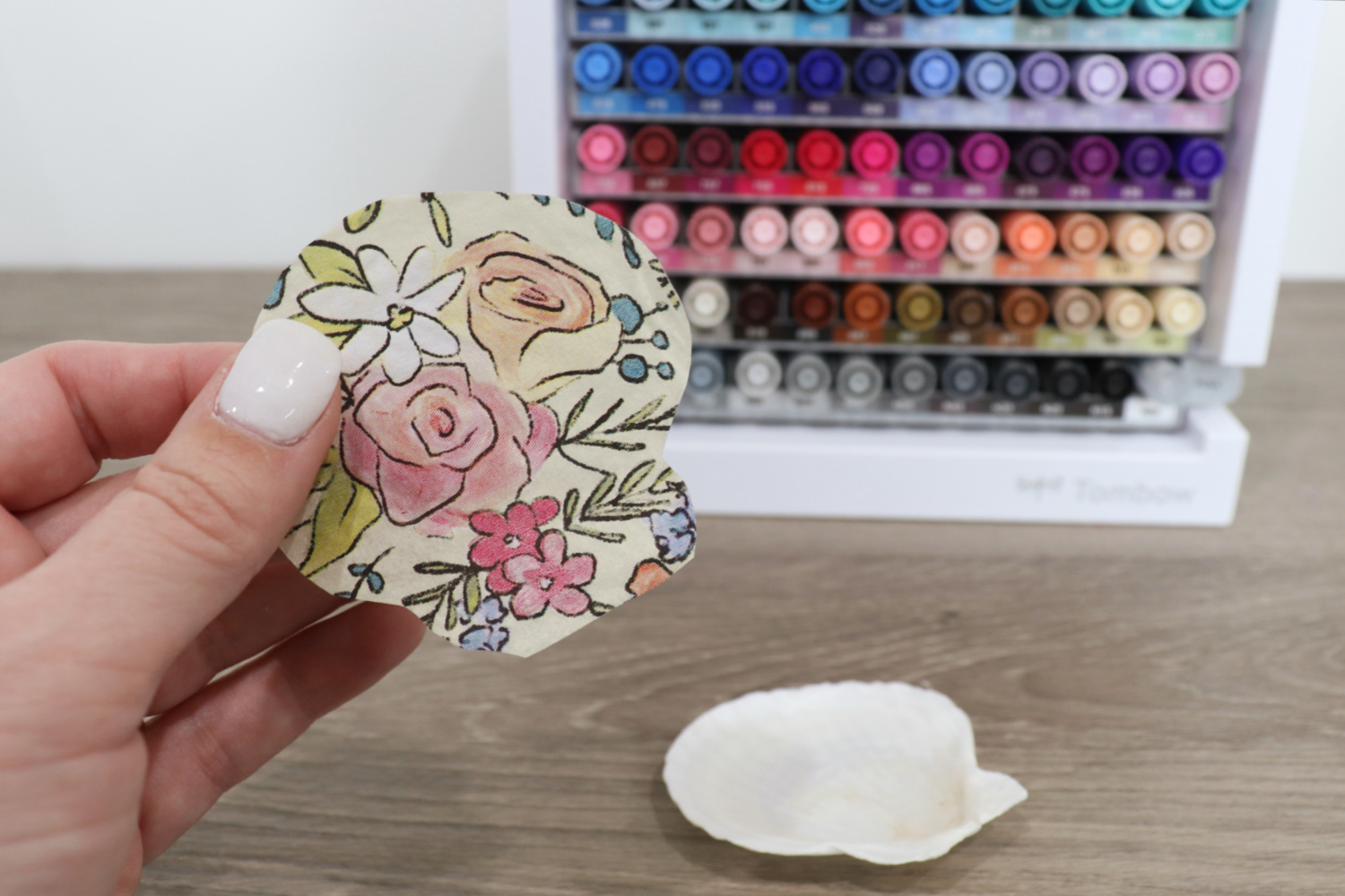 Image contains Amy’s hand holding a piece of patterned napkin cut in the shape of a scallop shell. The shell and a marker organizer sit nearby on a wooden table.