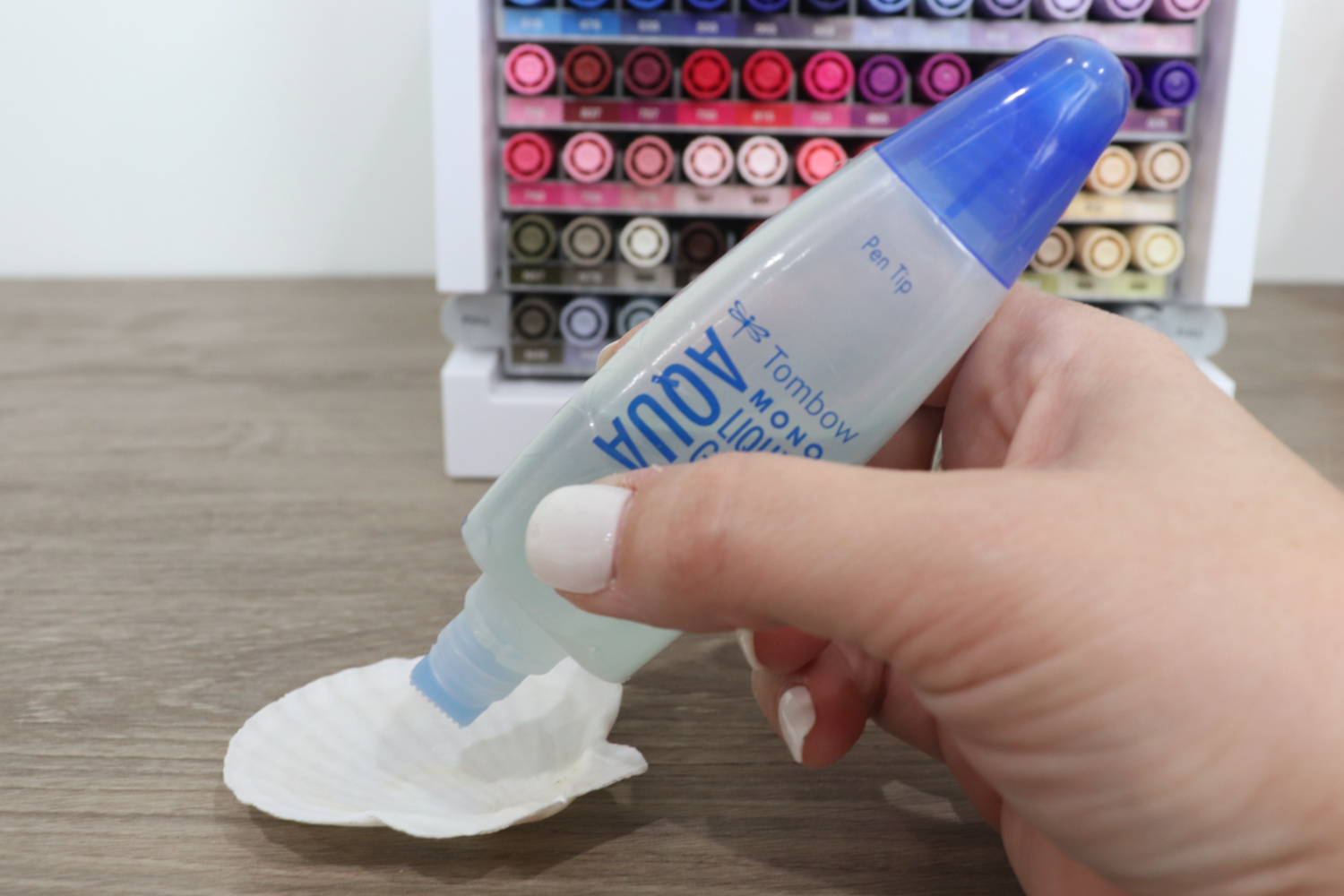 Image contains Amy’s hand holding a tube of Tombow MONO Liquid Glue and applying it to a white scalloped shell. A marker organizer sits nearby on a wooden desk.