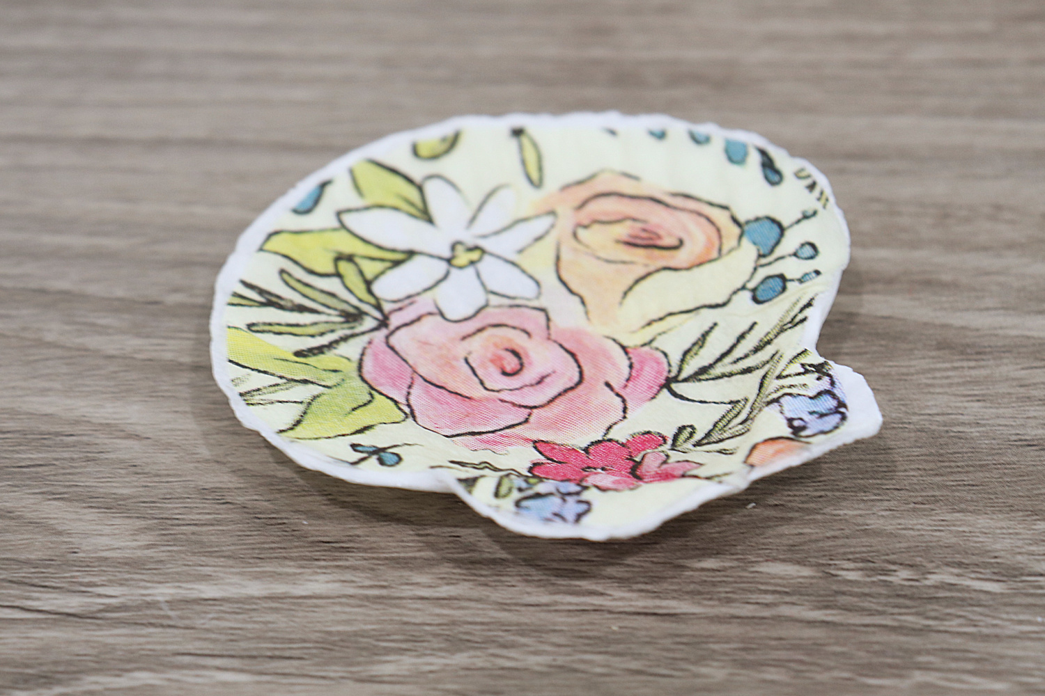 Image contains a scallop shell covered with a floral patterned napkin, sitting on a wooden desk.