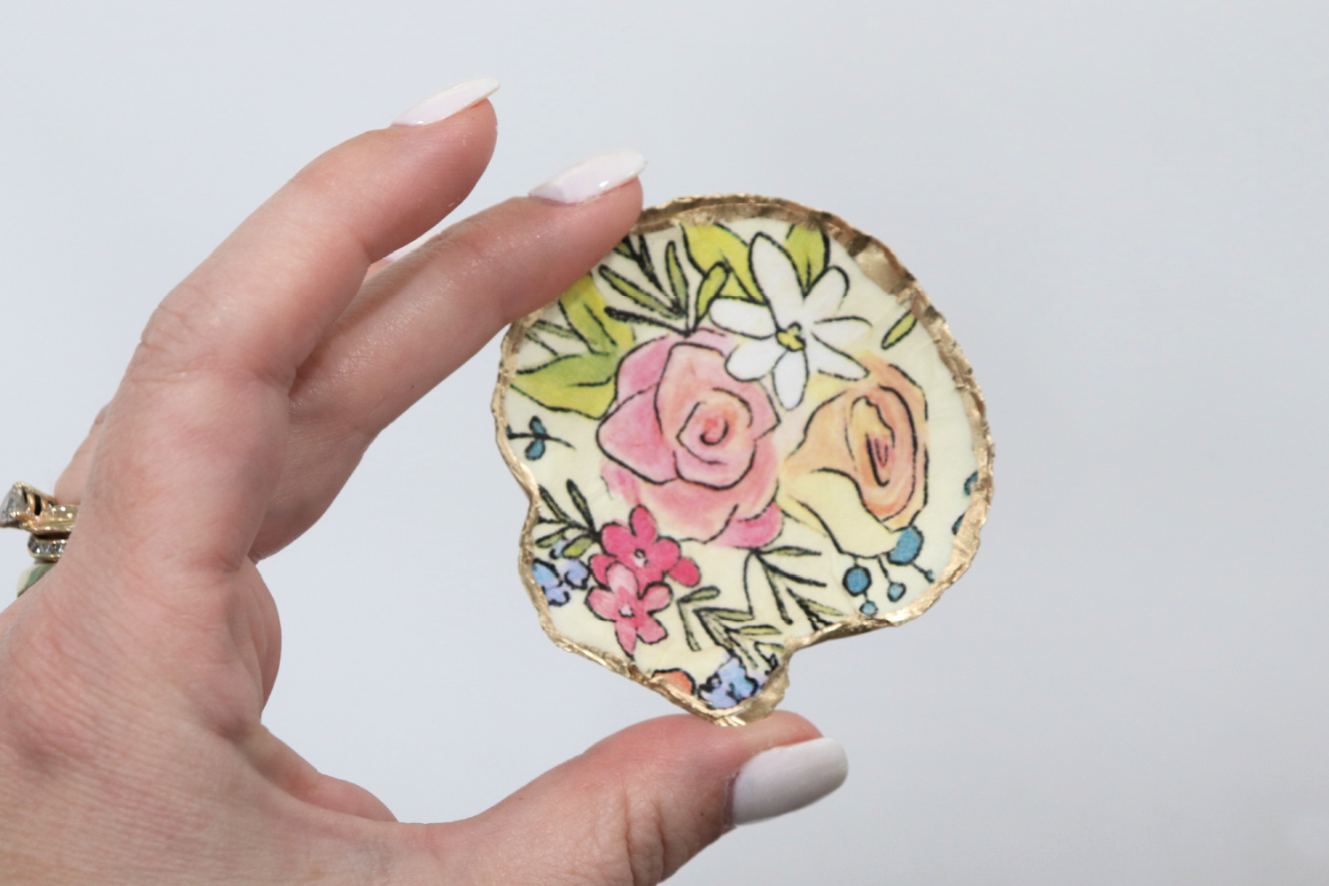 Image contains Amy’s hand holding a scallop shell jewelry dish. It is covered in a floral print and trimmed with gold paint.
