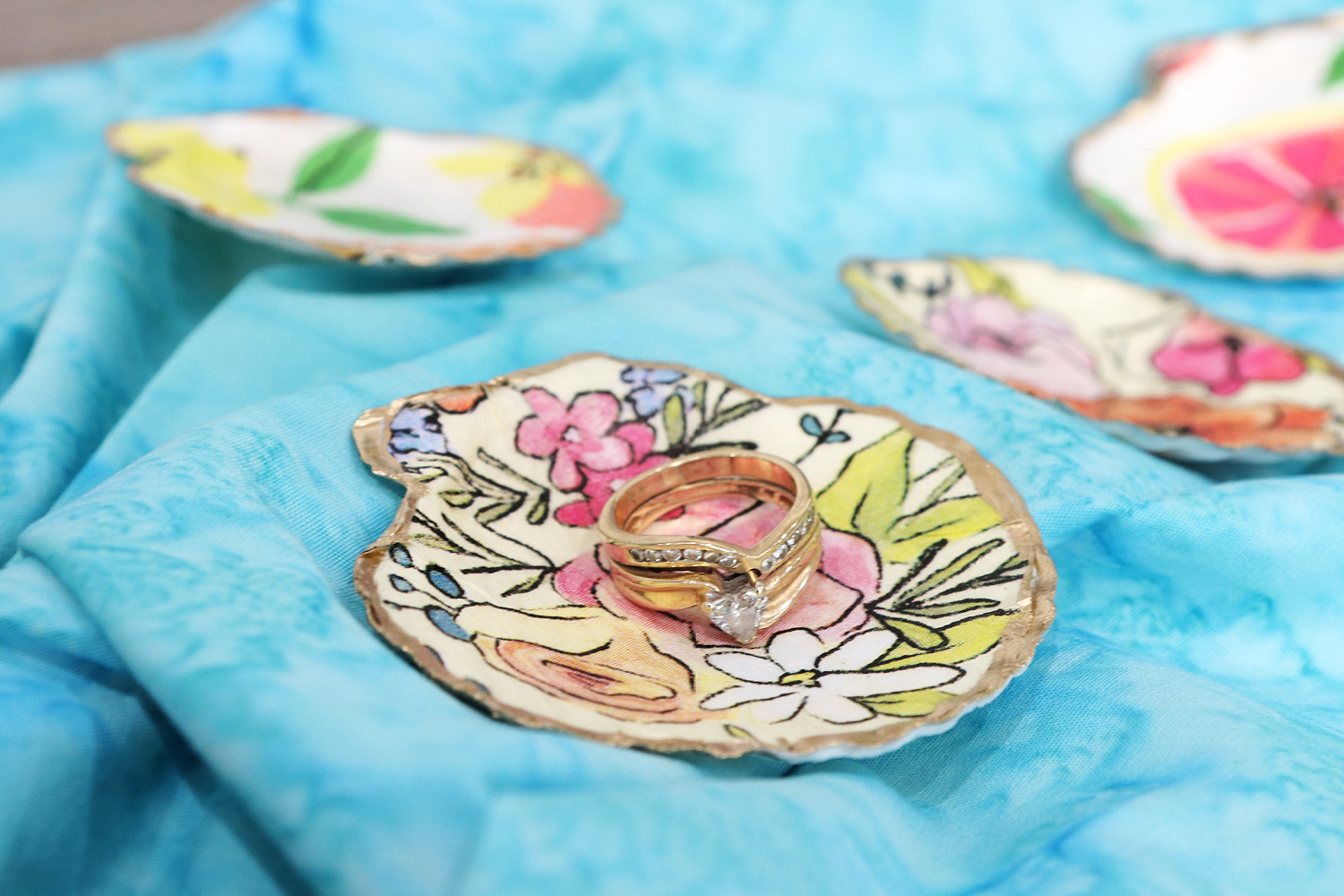 Image contains four decoupaged seashell jewelry dishes sitting on blue batik fabric. The one in focus holds a pair of gold and diamond rings.