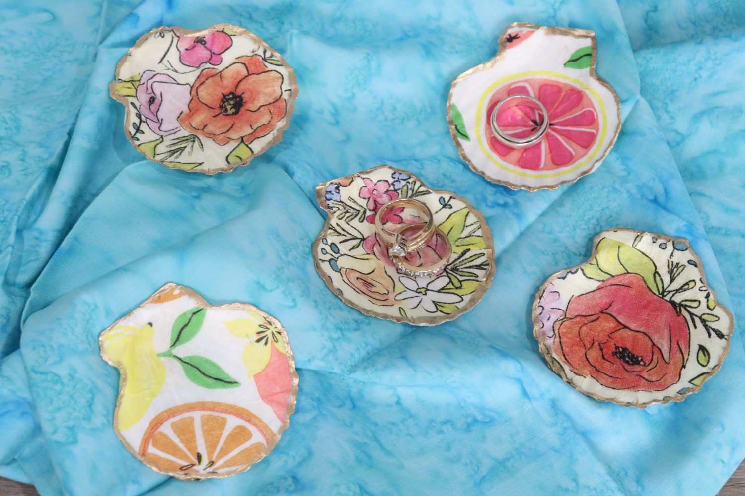 Image contains five decoupaged shell jewelry dishes arranged on a blue batik fabric background.