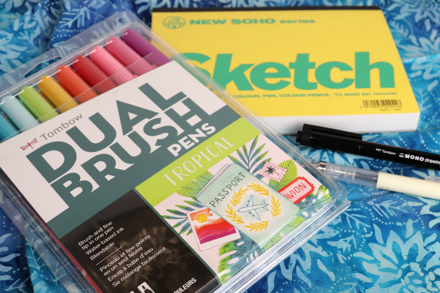 Image contains a pack of Dual Brush Pens in tropical colors, a yellow covered sketch pad, a MONO Drawing Pen, and a mechanical pencil on a blue batik fabric background.