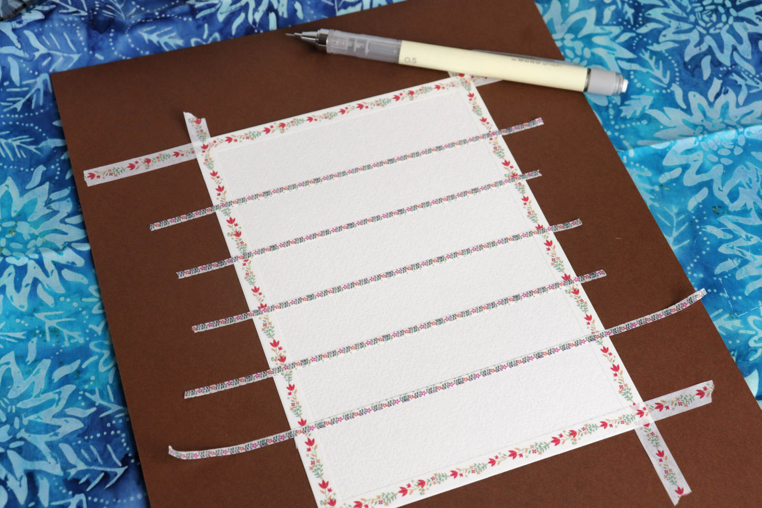 Image contains a white postcard sized paper attached to a piece of cardstock with washi tape. A mechanical pencil sits nearby. The postcard is divided into six horizontal stripes with 3mm thick washi tape.