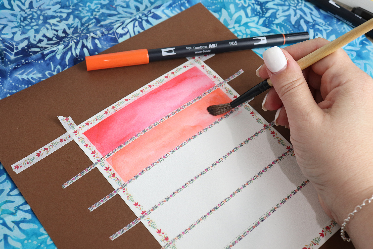 Image contains Amy’s hand holding a paintbrush and applying orange ink to a section of postcard. The top section has been watercolored red. An orange marker sits nearby.