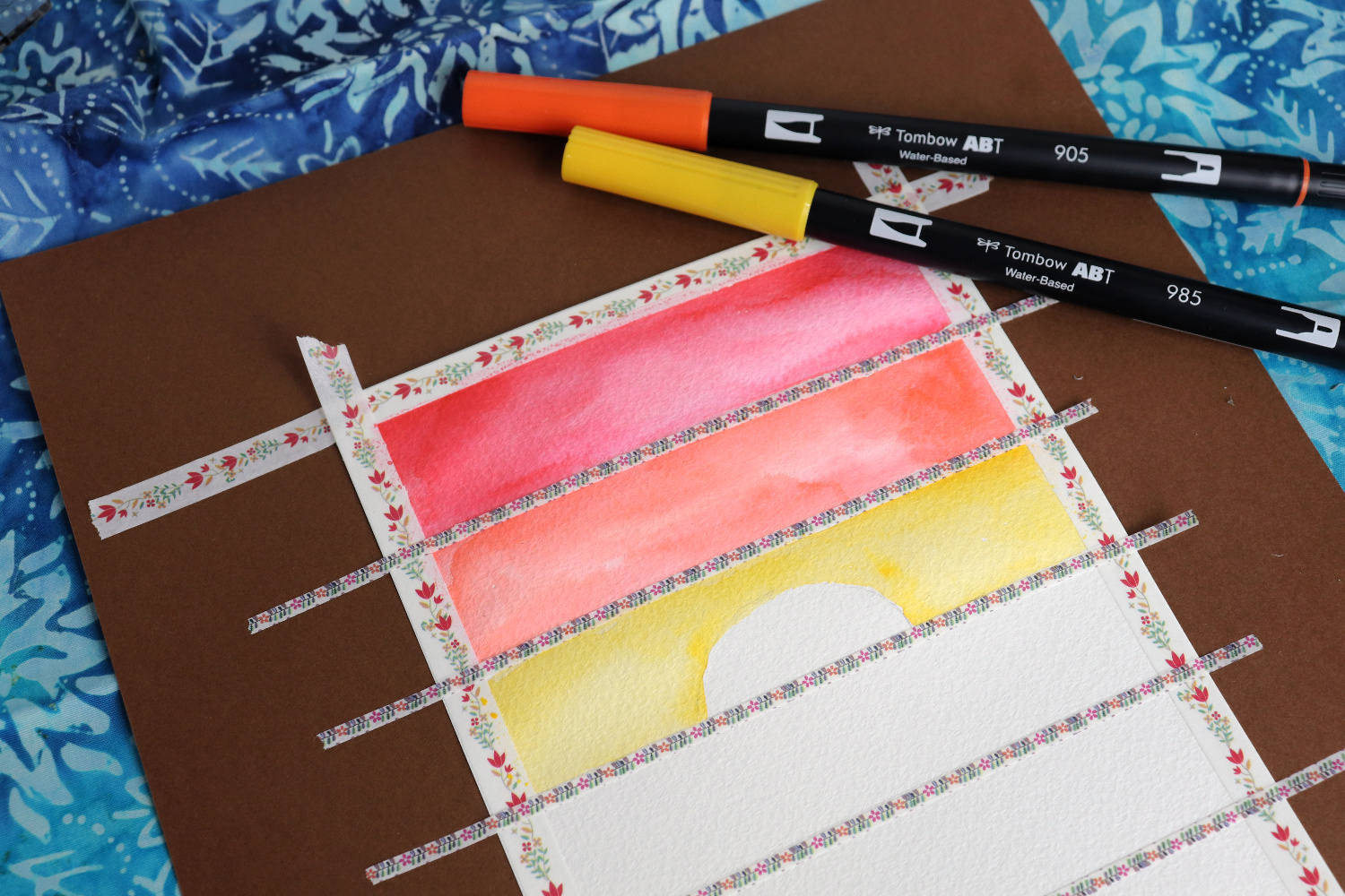 Image contains a white postcard divided into horizontal stripes with washi tape. The top two sections are colored reed and orange. The third is yellow with a white semi-circle left uncolored in the center. Orange and yellow markers sit nearby.