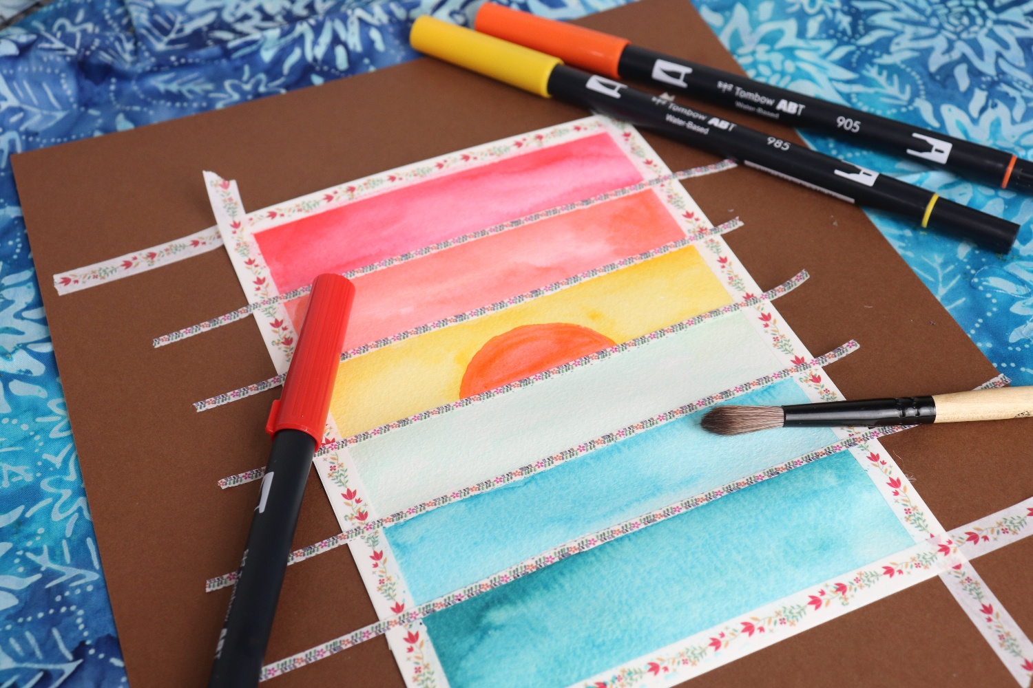 Image contains a postcard with horizontal stripes of watercolor in red, orange, yellow, and three shades of blue. Colored markers and a paintbrush lay nearby.