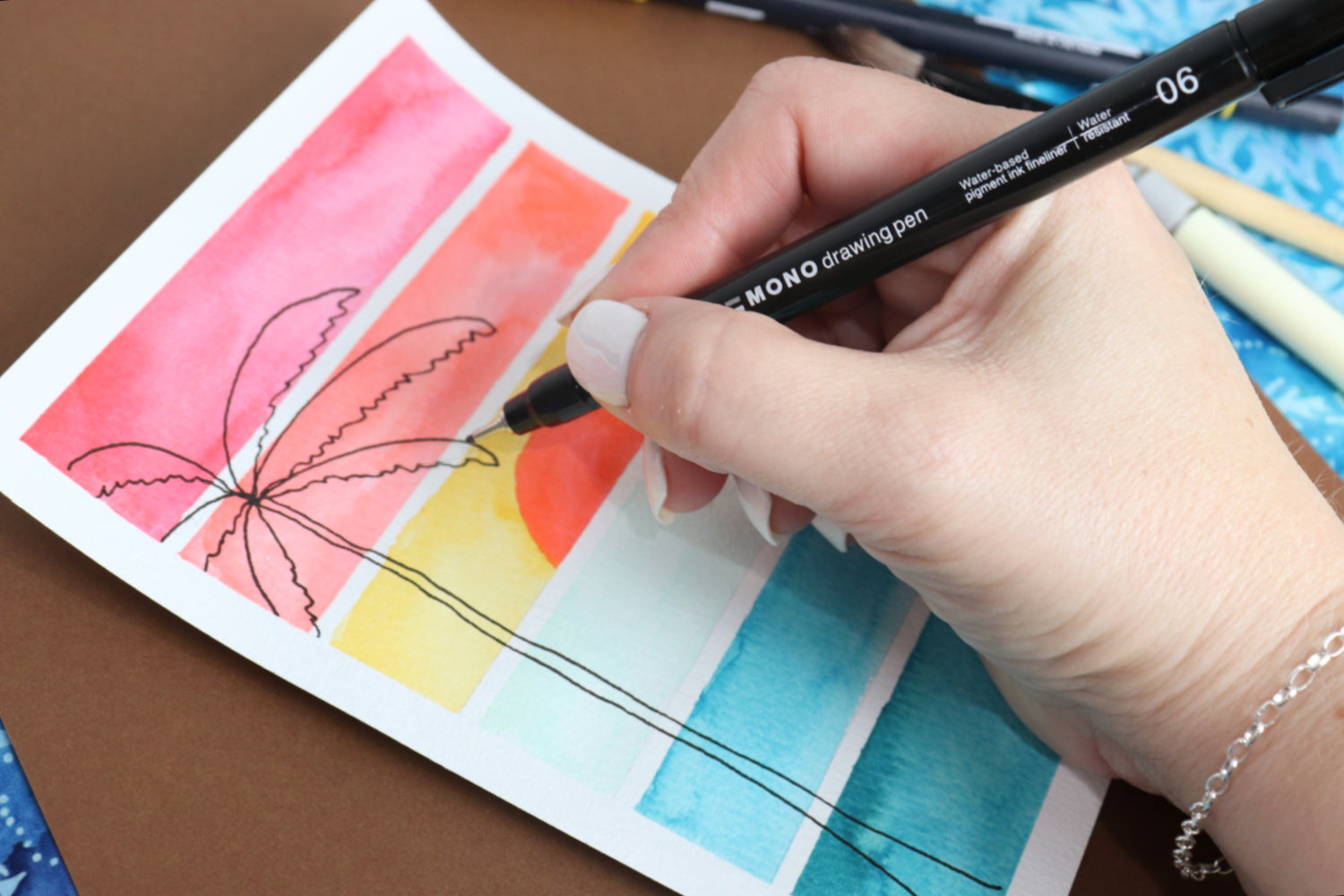 Image contains Amy’s hand holding a MONO Drawing Pen and sketching the outline of a palm tree on the watercolored postcard.