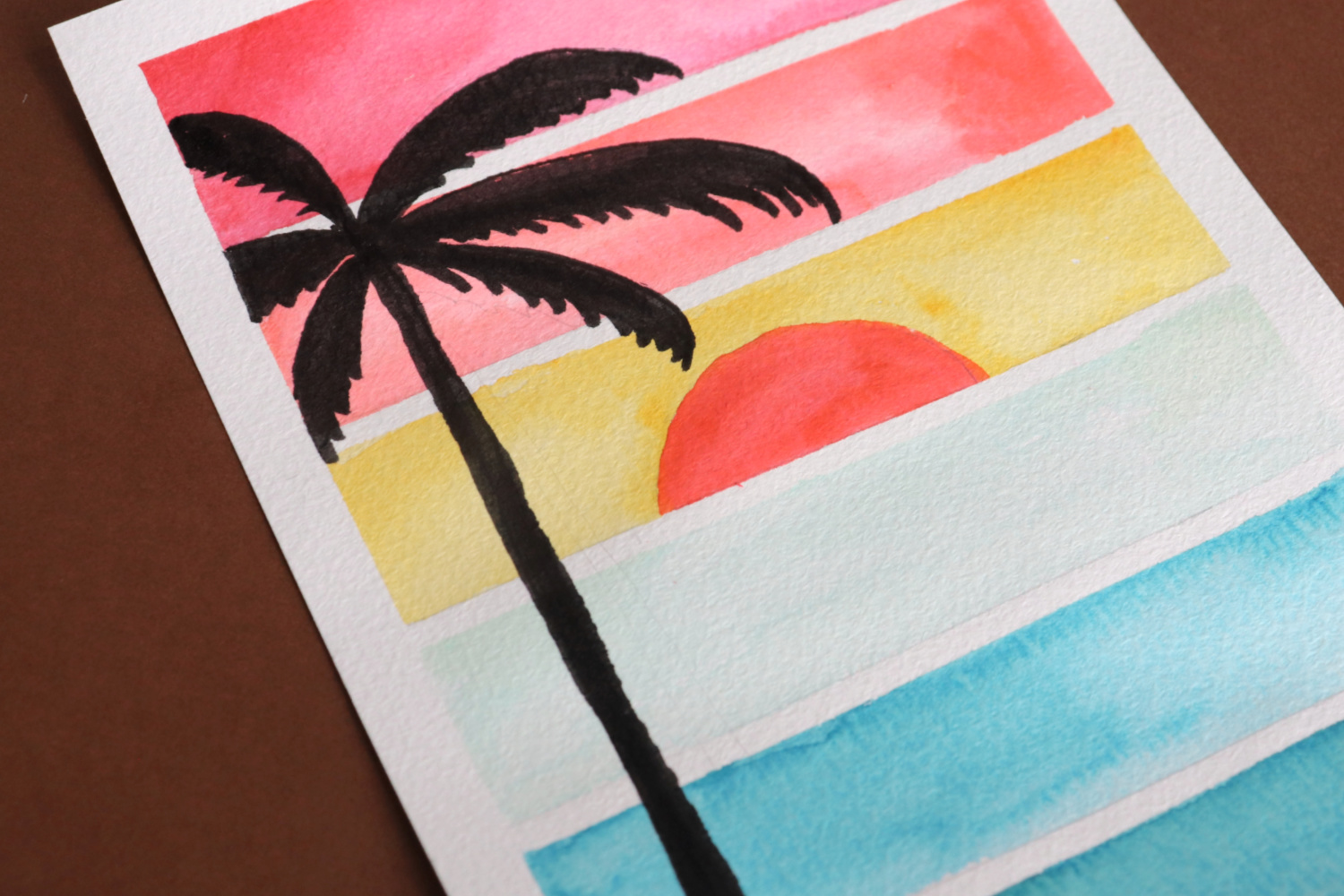 Image contains a postcard that has six watercolored stripes: red, orange, yellow, and three shades of blue. An orange semi-circle sun sits in the center of the yellow stripe, and the silhouette of a palm tree goes down the left side.