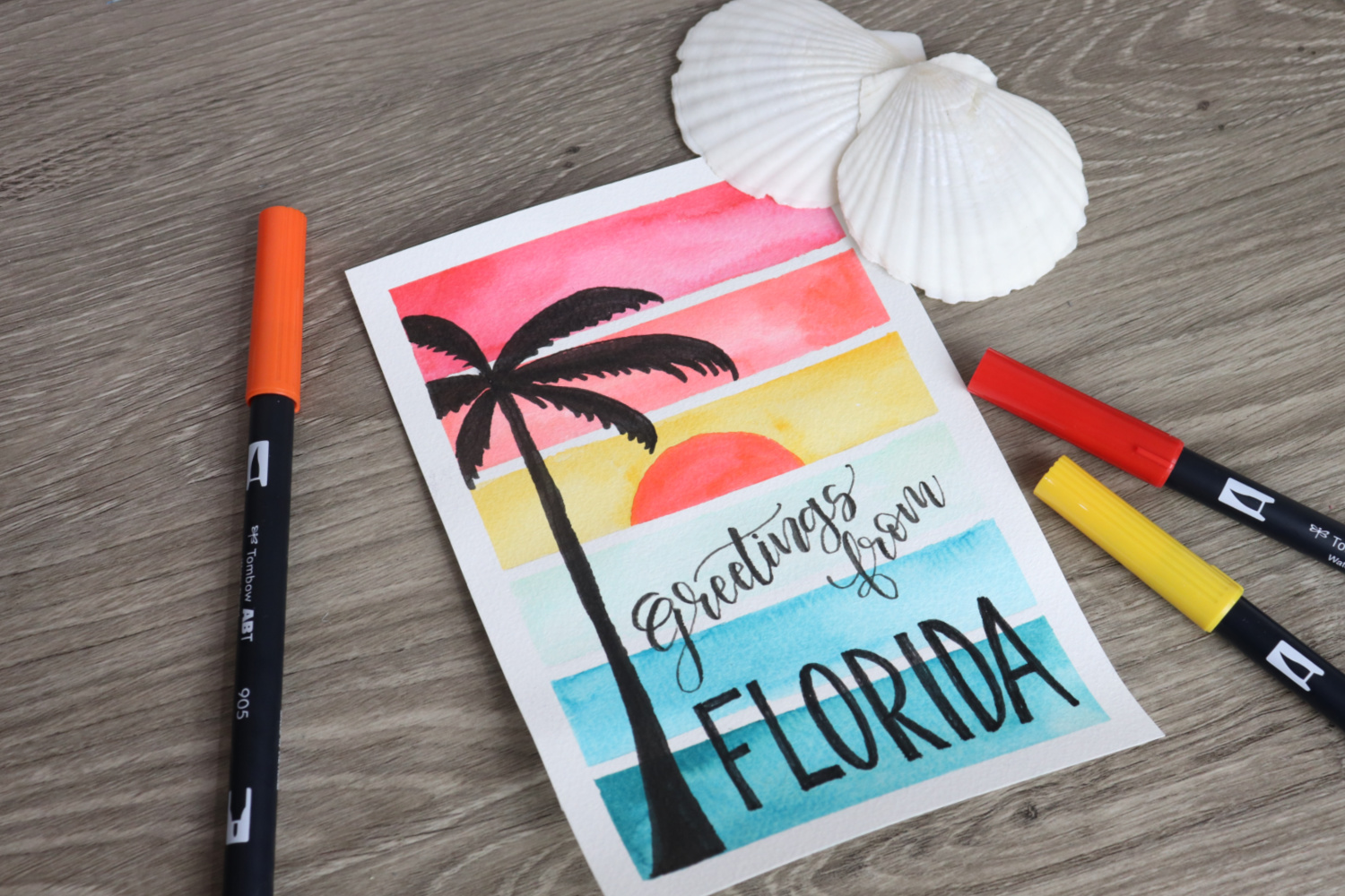 Image contains a watercolor sunset postcard with a palm tree silhouette and the words, “Greetings from Florida.” Three markers and two scallop shells lay around it on a wooden desktop.