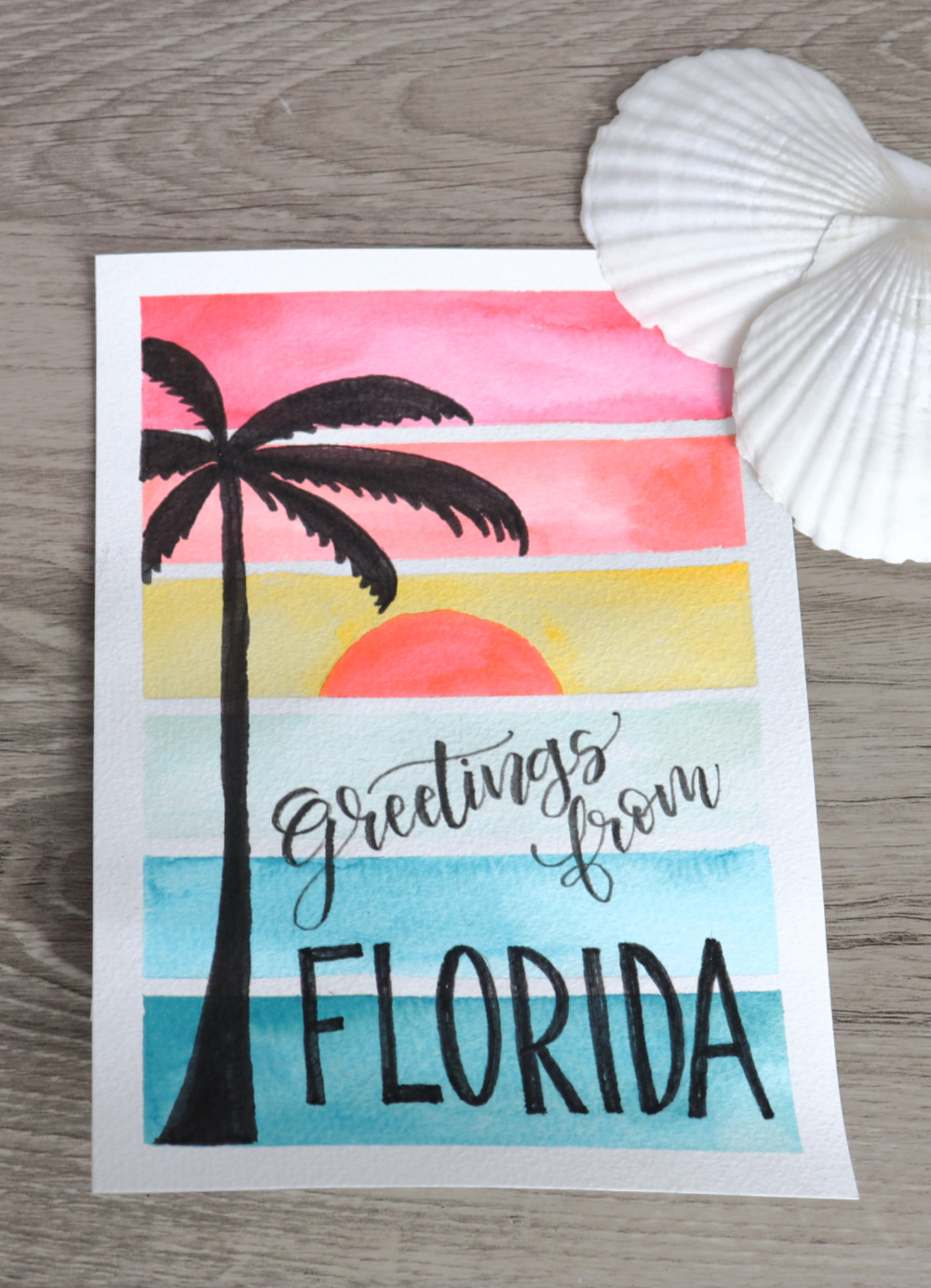Image contains a watercolor sunset postcard with a palm tree and the words, “Greetings from Florida.” A pair of scallop shells sit off to the side on a wooden desk.