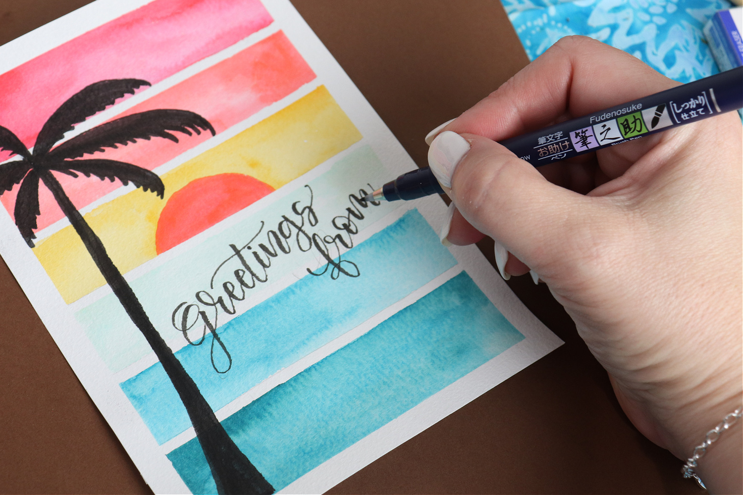 Image contains Amy’s hand using a Tombow Fudenosuke to letter the words “greetings from” on a watercolor sunset postcard.