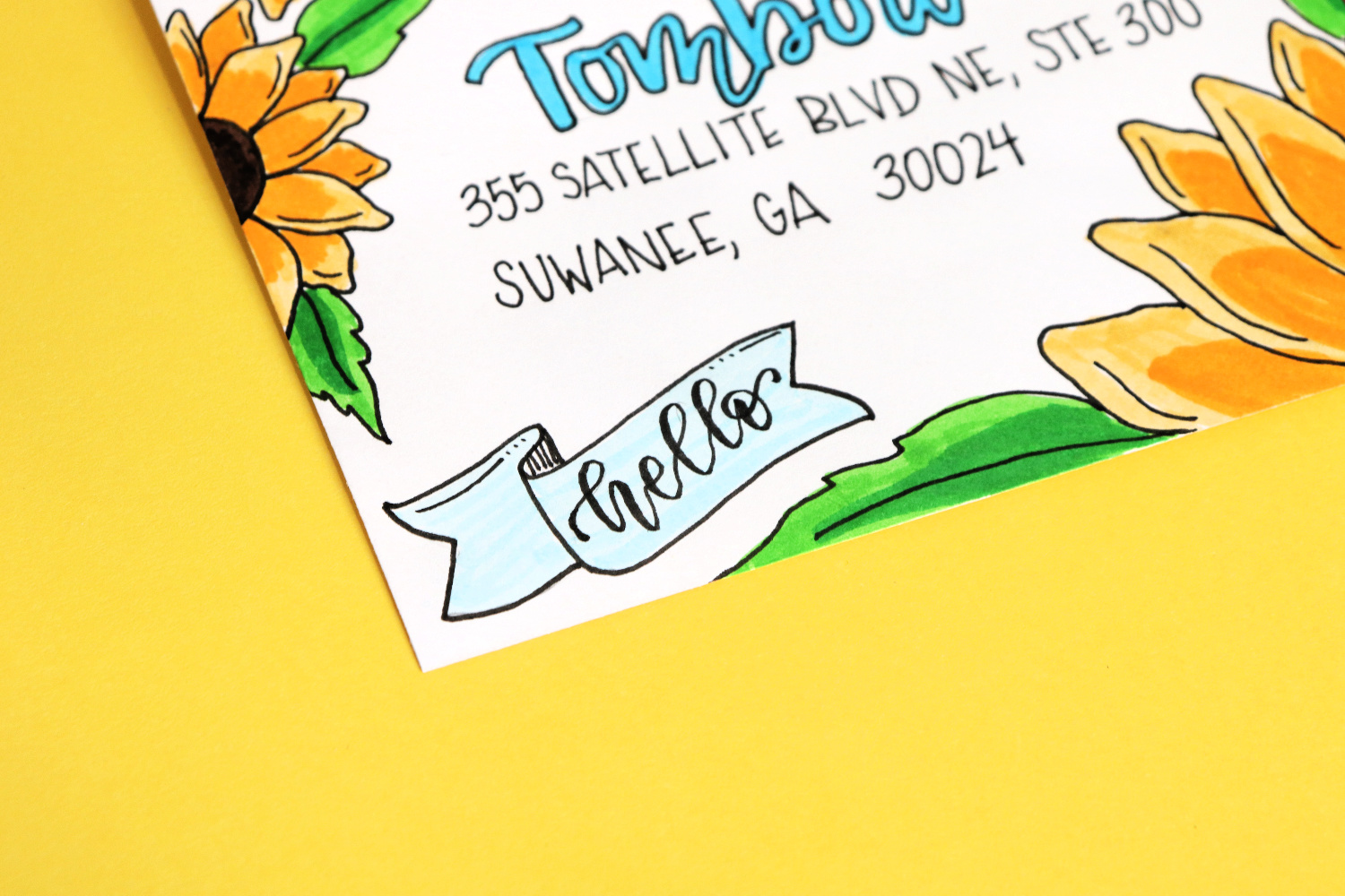 Image contains a close-up view of the bottom left corner of a white sunflower envelope. A blue banner is in the corner with the word “hello” hand lettered on it. 