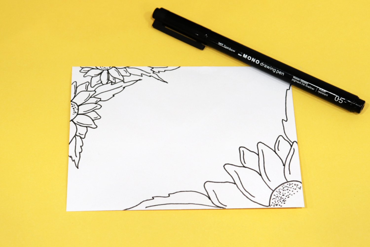 Image contains a white envelope with three partial sunflowers drawn around the edges in black ink. A black MONO Drawing Pen sits nearby on a yellow background.