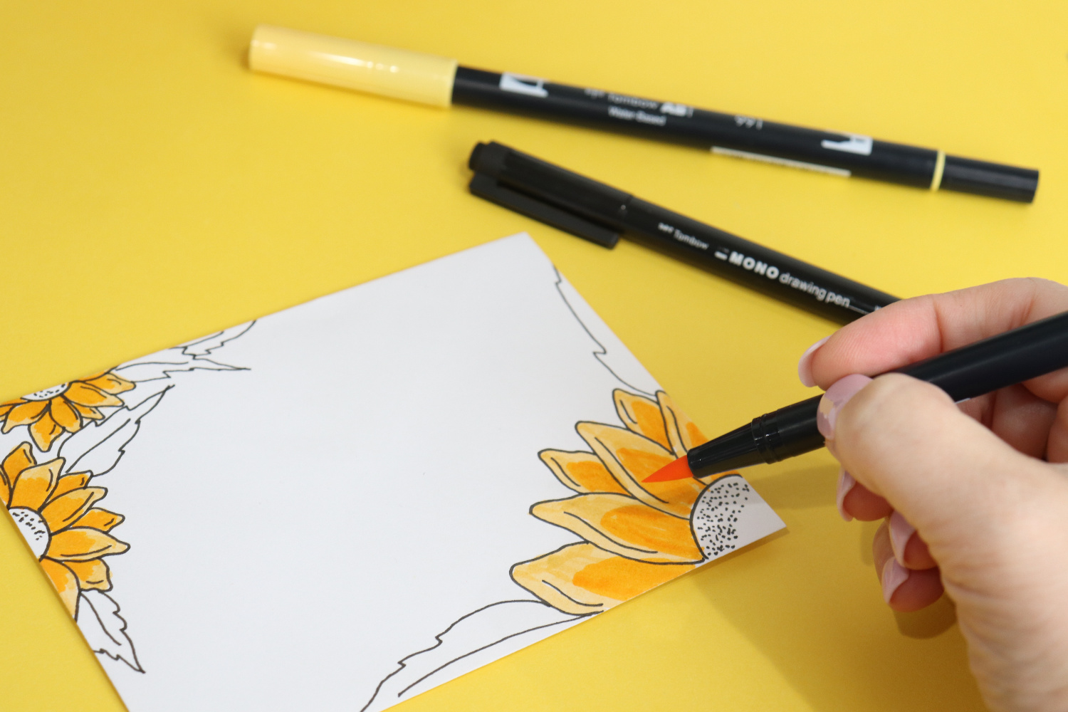 Image contains Amy’s hand holding a yellow Dual Brush Pen and coloring in the sunflower drawings on a white envelope. Two other markers lay nearby on a yellow background.