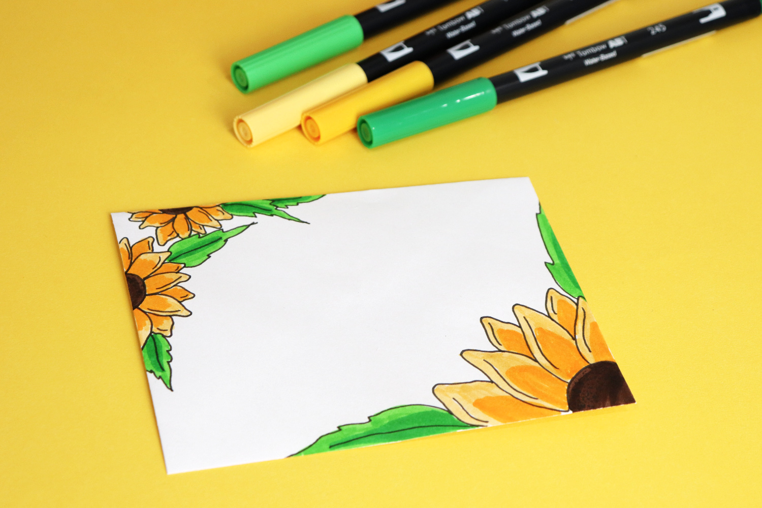 Image contains a white envelope decorated with colored sunflowers laying on a yellow background. Two yellow and two green markers lay nearby.