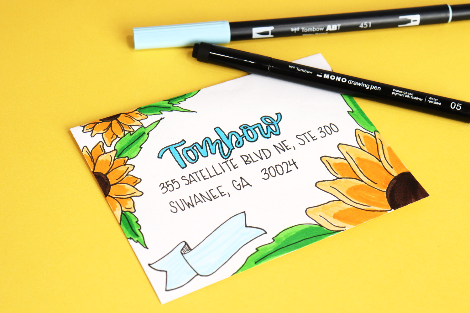 Image contains a white envelope decorated with yellow sunflowers and addressed to Tombow’s Georgia office. A blue banner is drawn in the lower left corner. Two markers sit nearby, all on a yellow background.