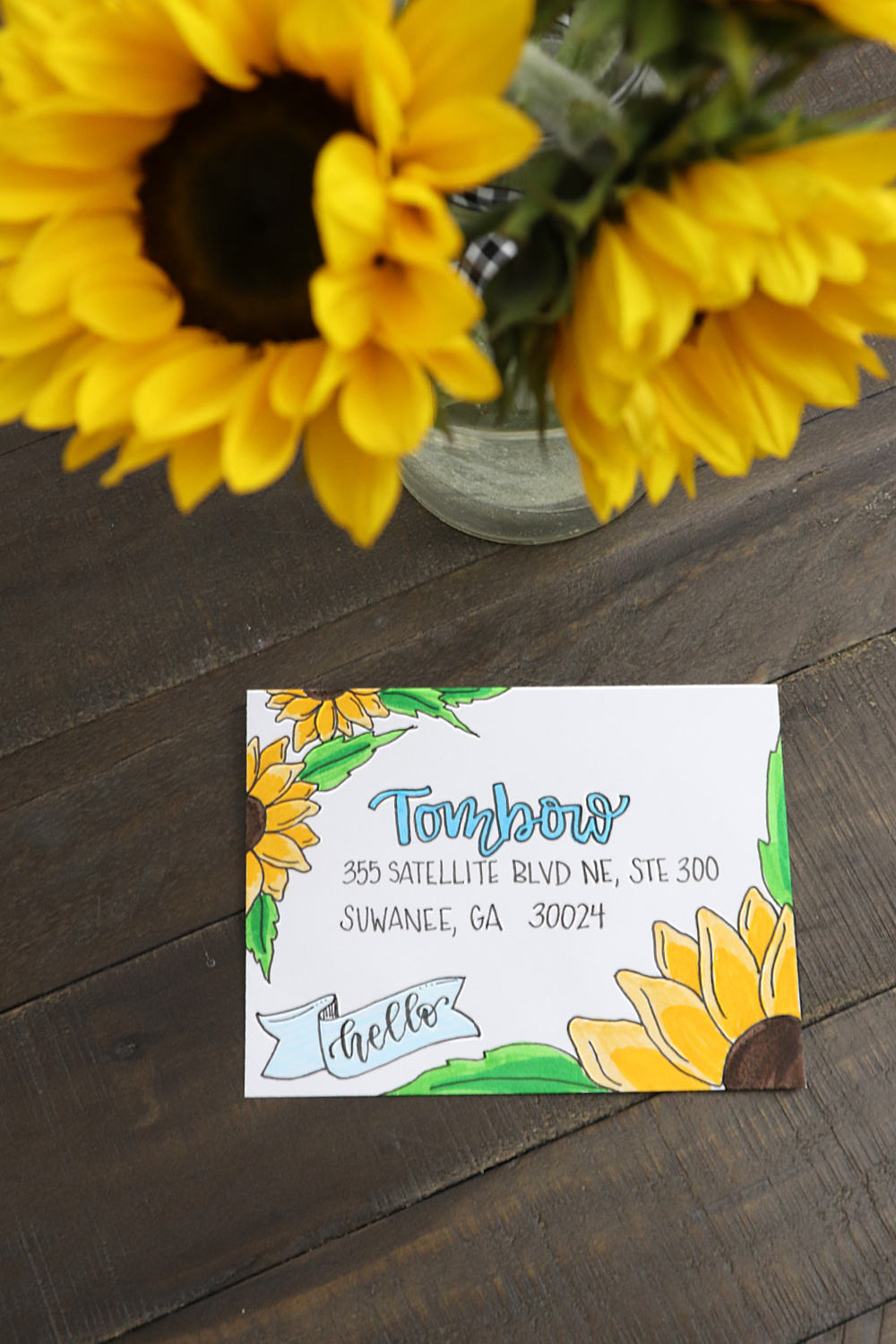 Image contains a white envelope decorated with sunflowers. A vase of sunflowers sits nearby on a dark farmhouse table top.