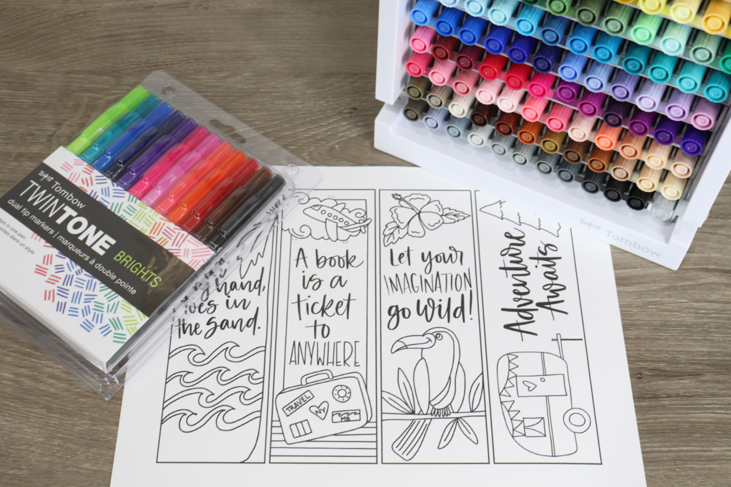 Image contains a sheet of white cardstock with four summer adventure bookmarks printed on it. A marker organizer filled with Dual Brush Pens, and a pack of TwinTone Brights sit nearby on a wooden desktop.