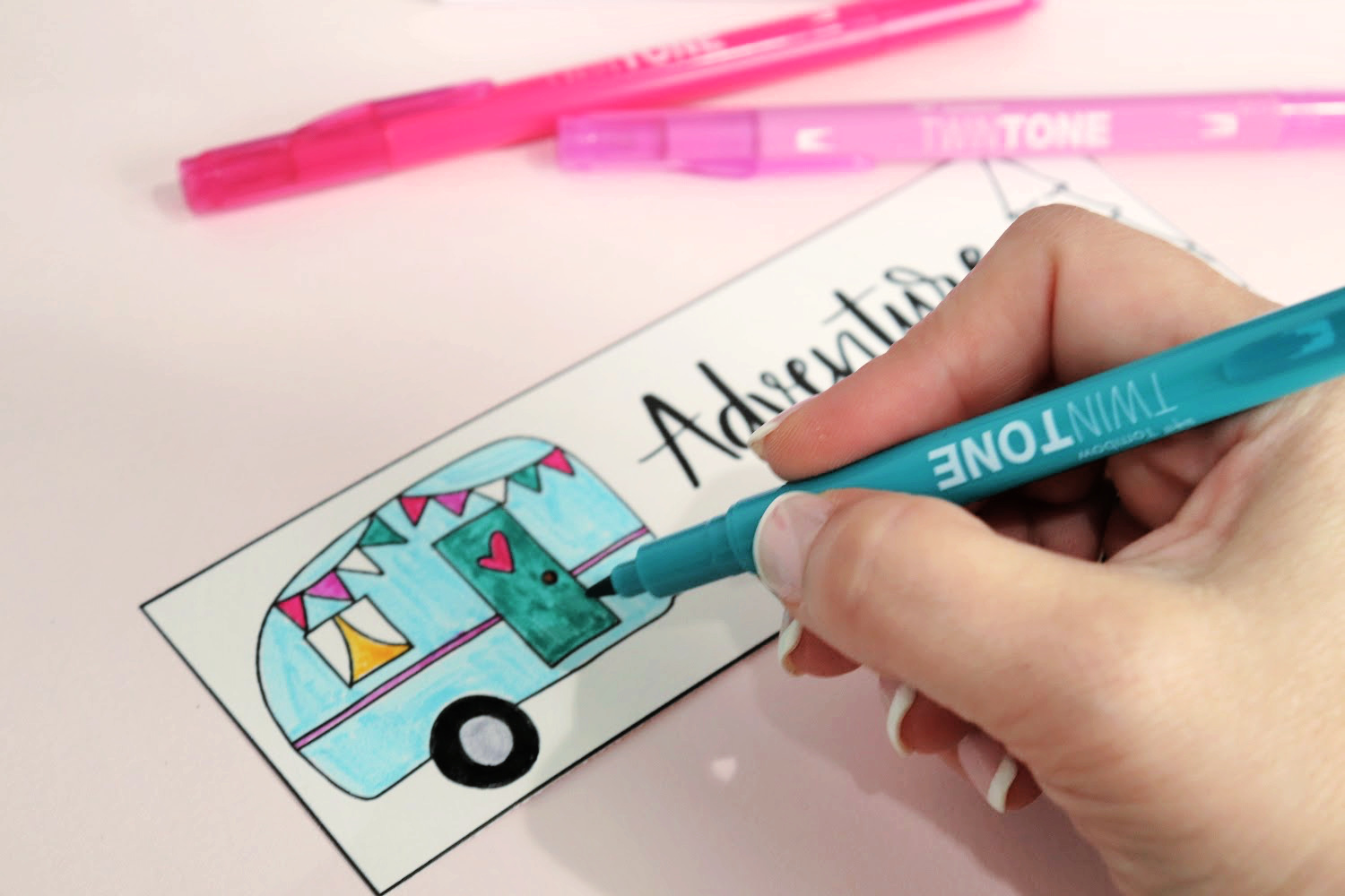 Image contains Amy’s hand holding a teal TwinTone marker, coloring a bookmark with a camper doodle. It sits on a pink background with two pink markers nearby.