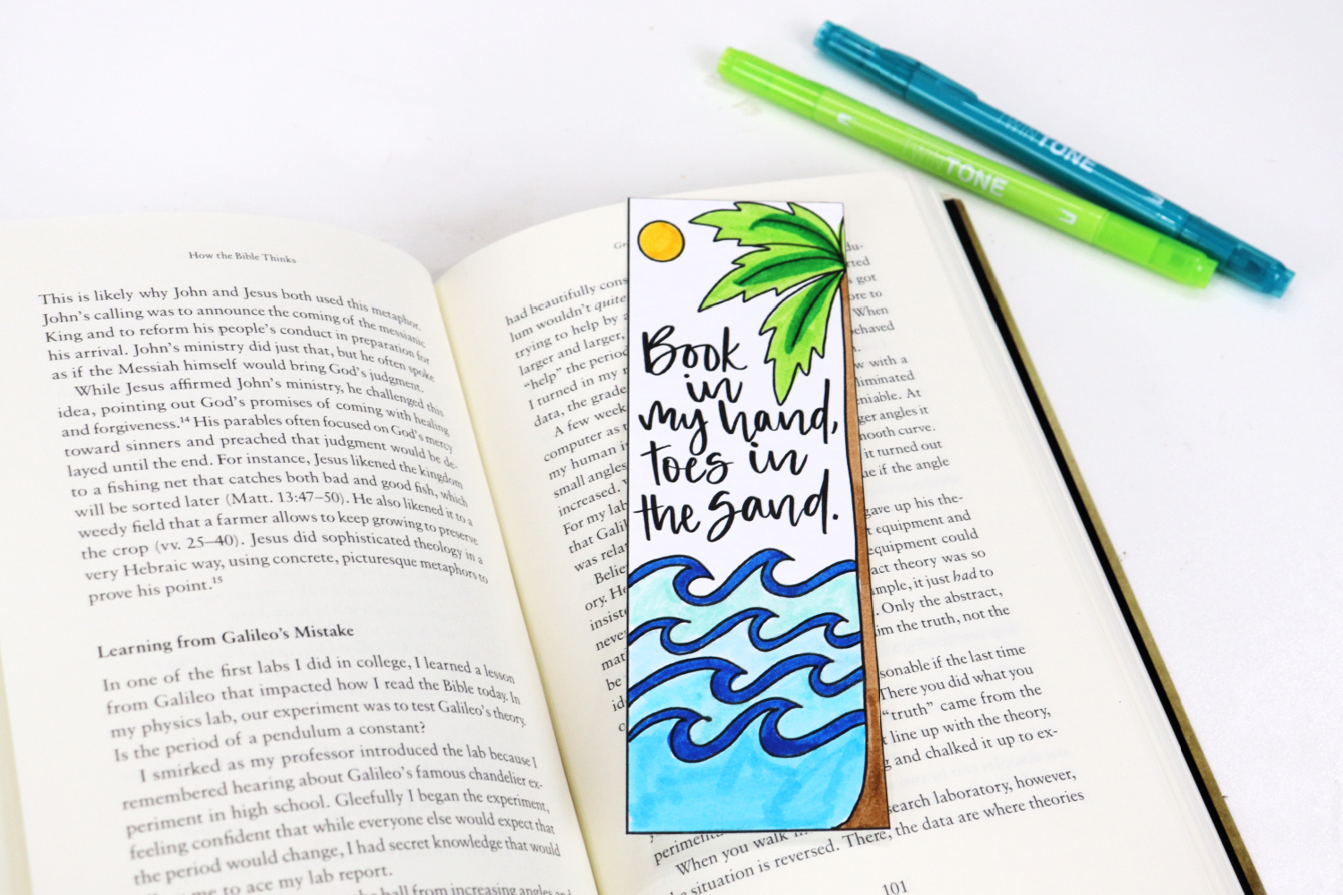 Image contains an open book with a beach themed summer bookmark inside. Blue and green markers sit nearby on a white background.
