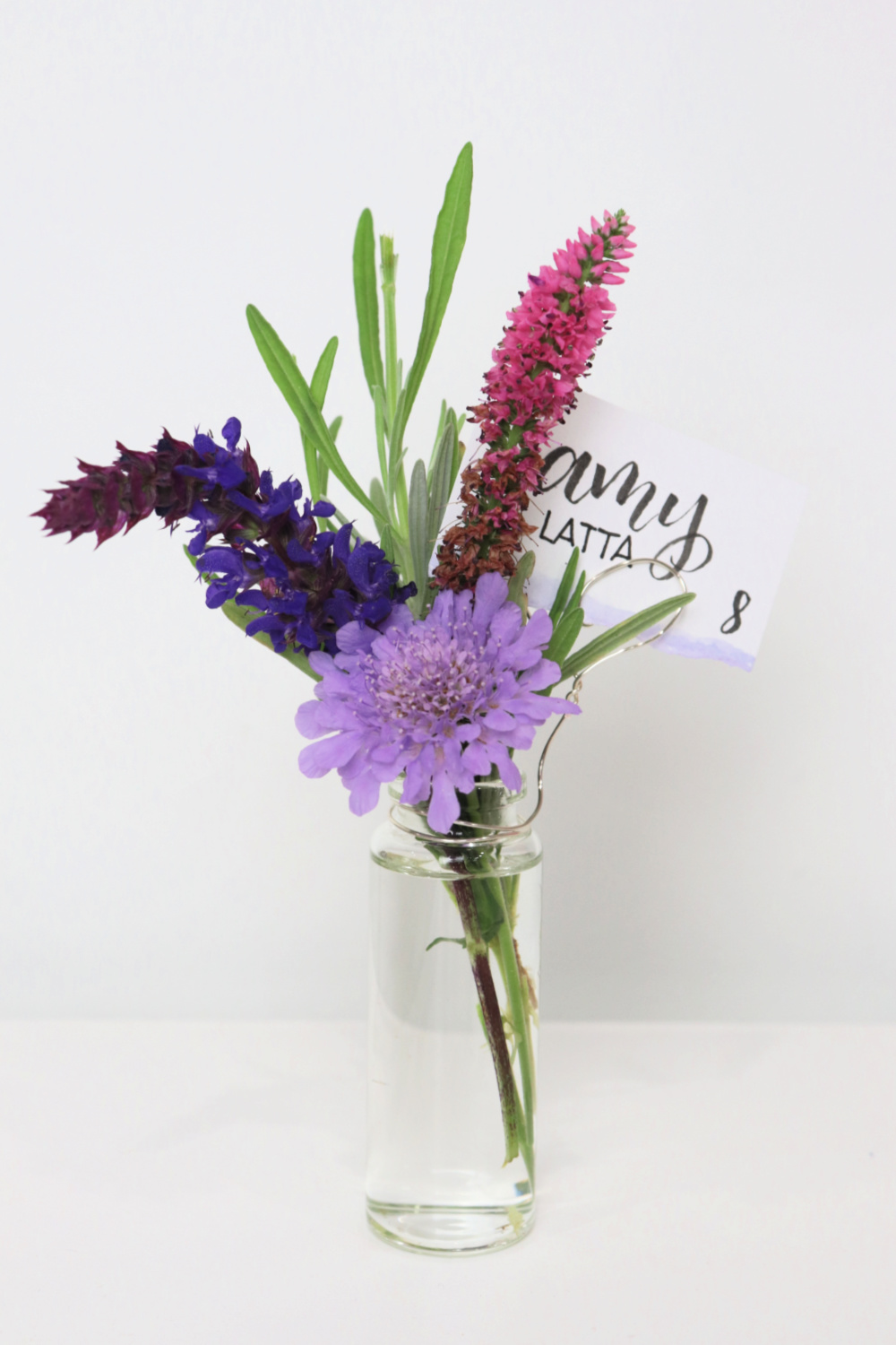 Image contains three small wildflowers and a sprig of lavender in a tiny jar. A piece of wire attached to the jar holds a cardstock place card with the words “Amy Latta” and table number “8”.