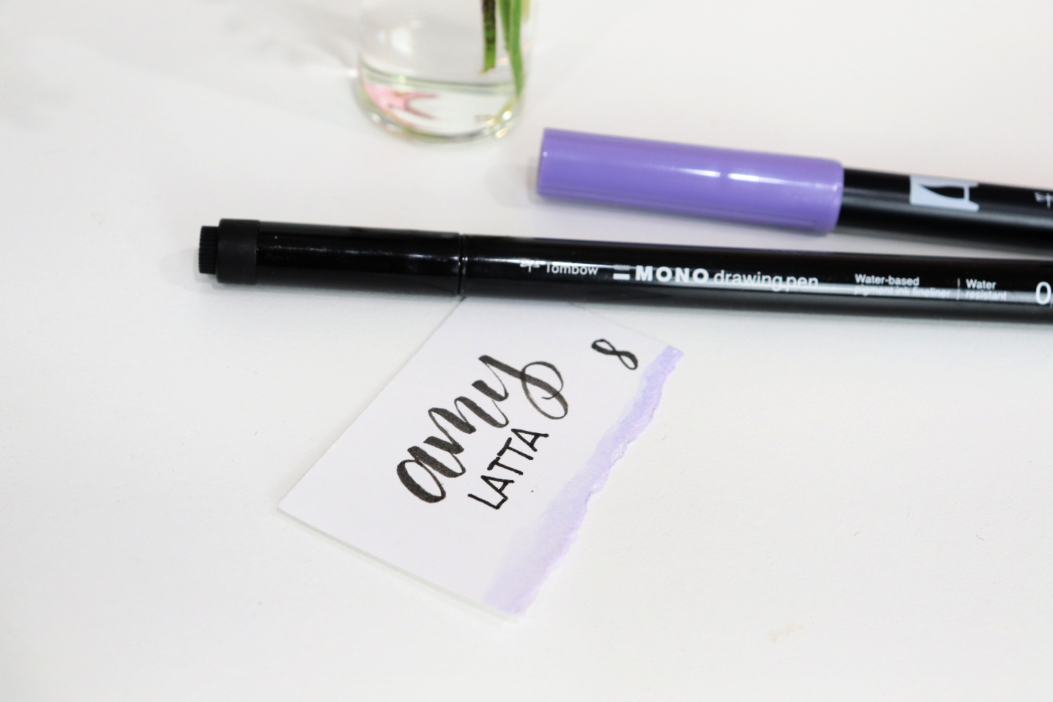 Image contains a small cardstock rectangle with the words “amy latta” and the number “8” written in black ink. The bottom of the card is painted light purple. A black marker and a purple marker lay nearby.
