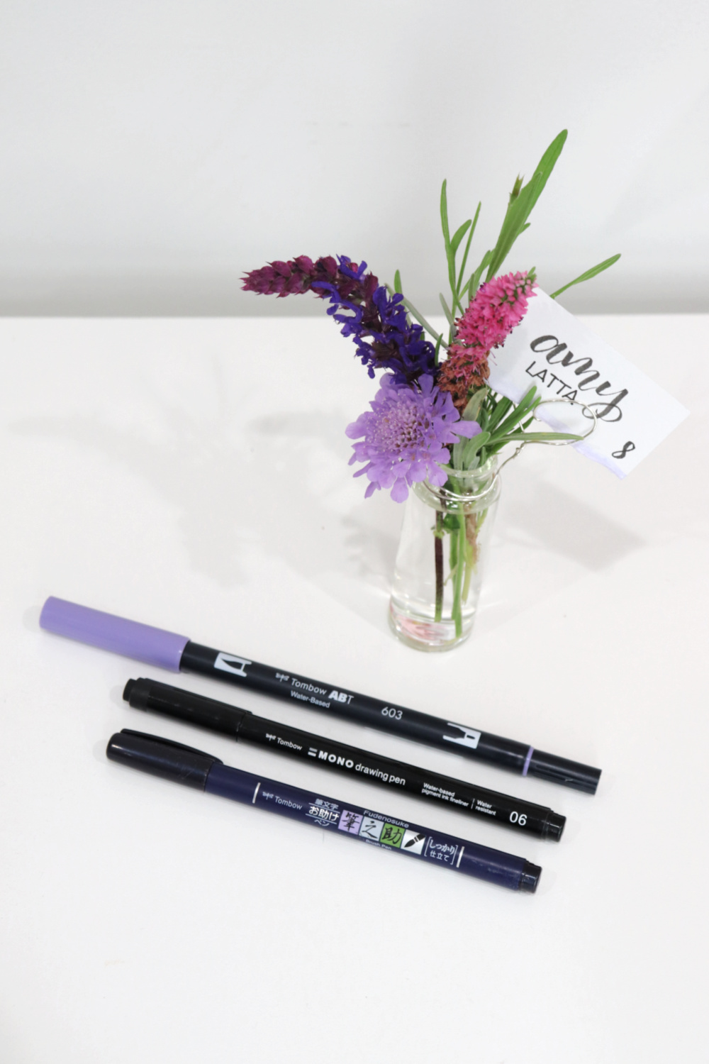 Image contains three small wildflowers and a sprig of lavender in a tiny jar. A piece of wire attached to the jar holds a cardstock place card with the words “Amy Latta” and table number “8”. Three markers lay nearby.