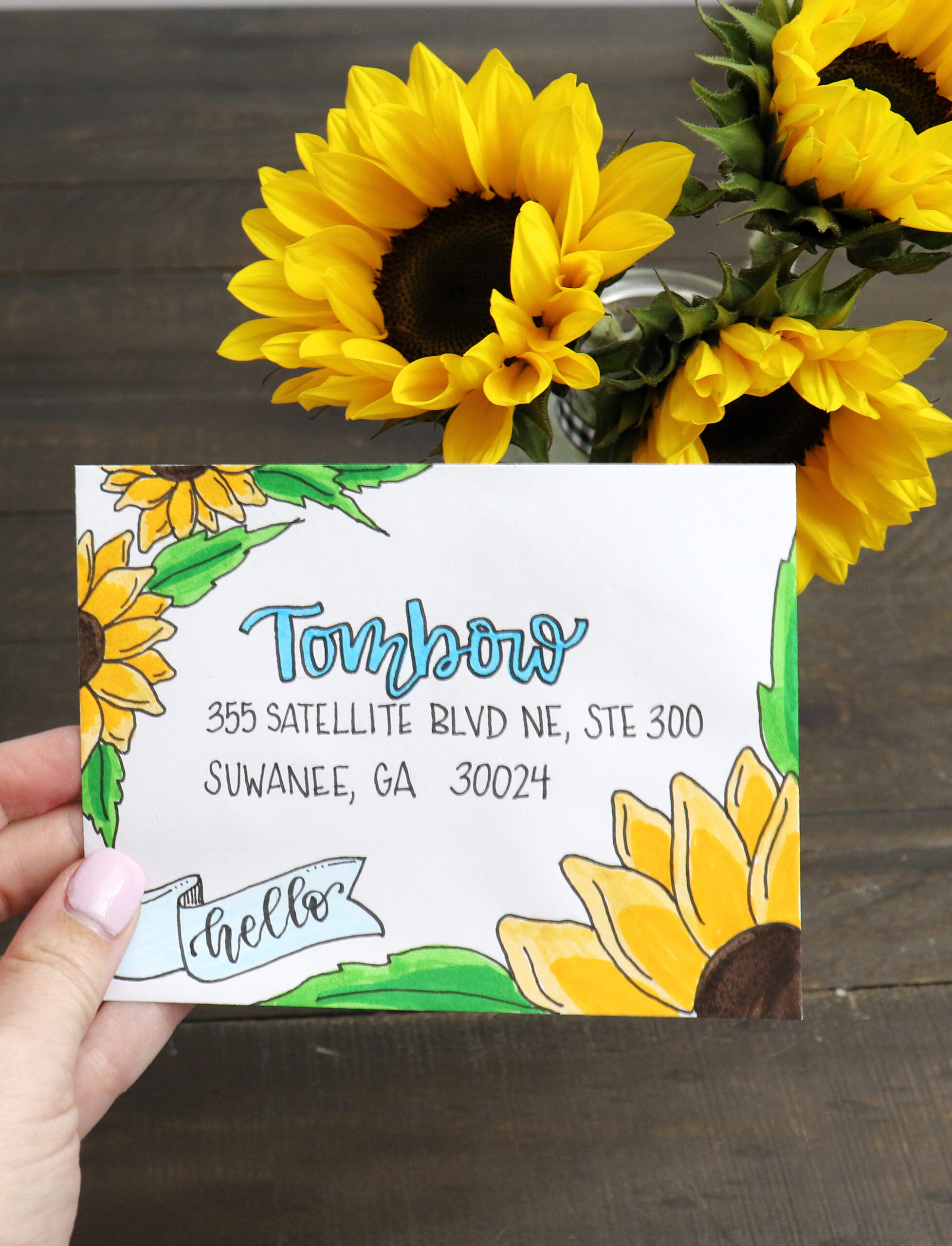 Image contains Amy’s hand holding a white envelope decorated with sunflowers. A vase of sunflowers sits in the background.