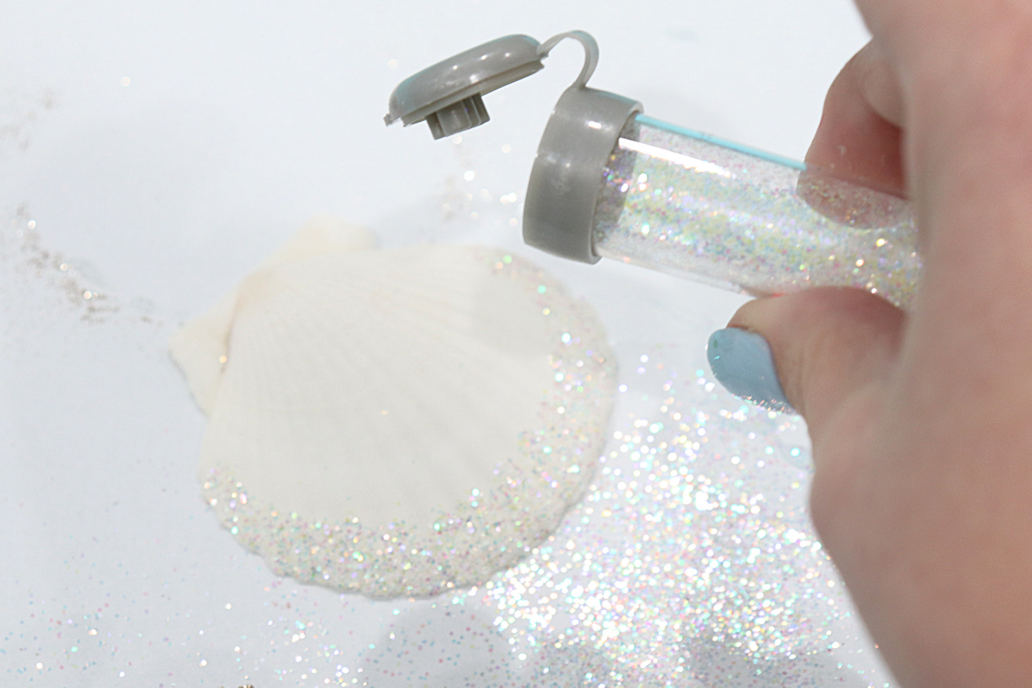 Image contains Amy’s hand holding a tube of iridescent white glitter above a scalloped seashell on a white table.