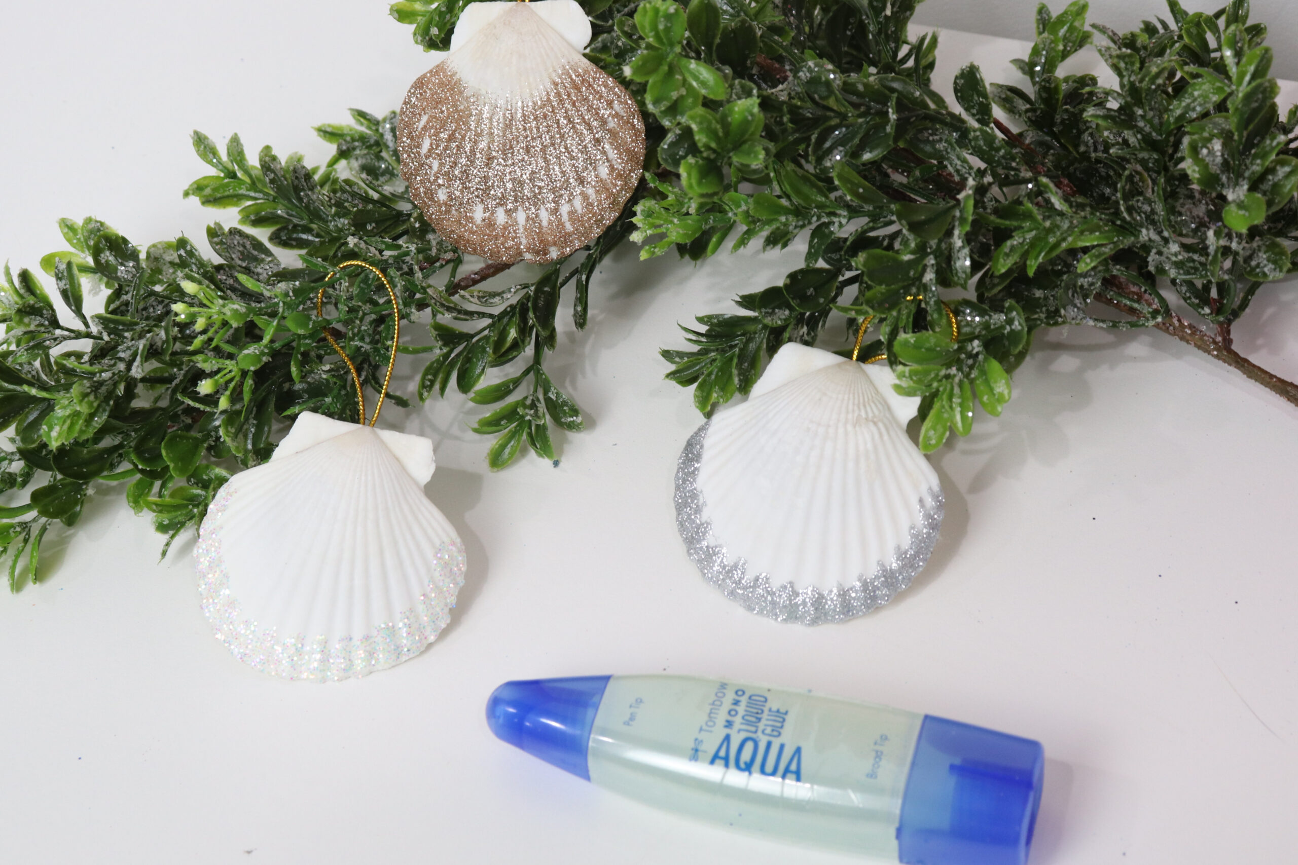 Image includes three glittered seashell ornaments lying on a faux branch on top of a white table. A tube of Tombow MONO Aqua Liquid Glue sits nearby.