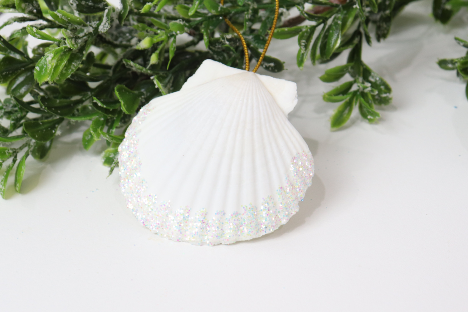 Image contains a white seashell ornament accented with iridescent white glitter laying near a faux green branch.