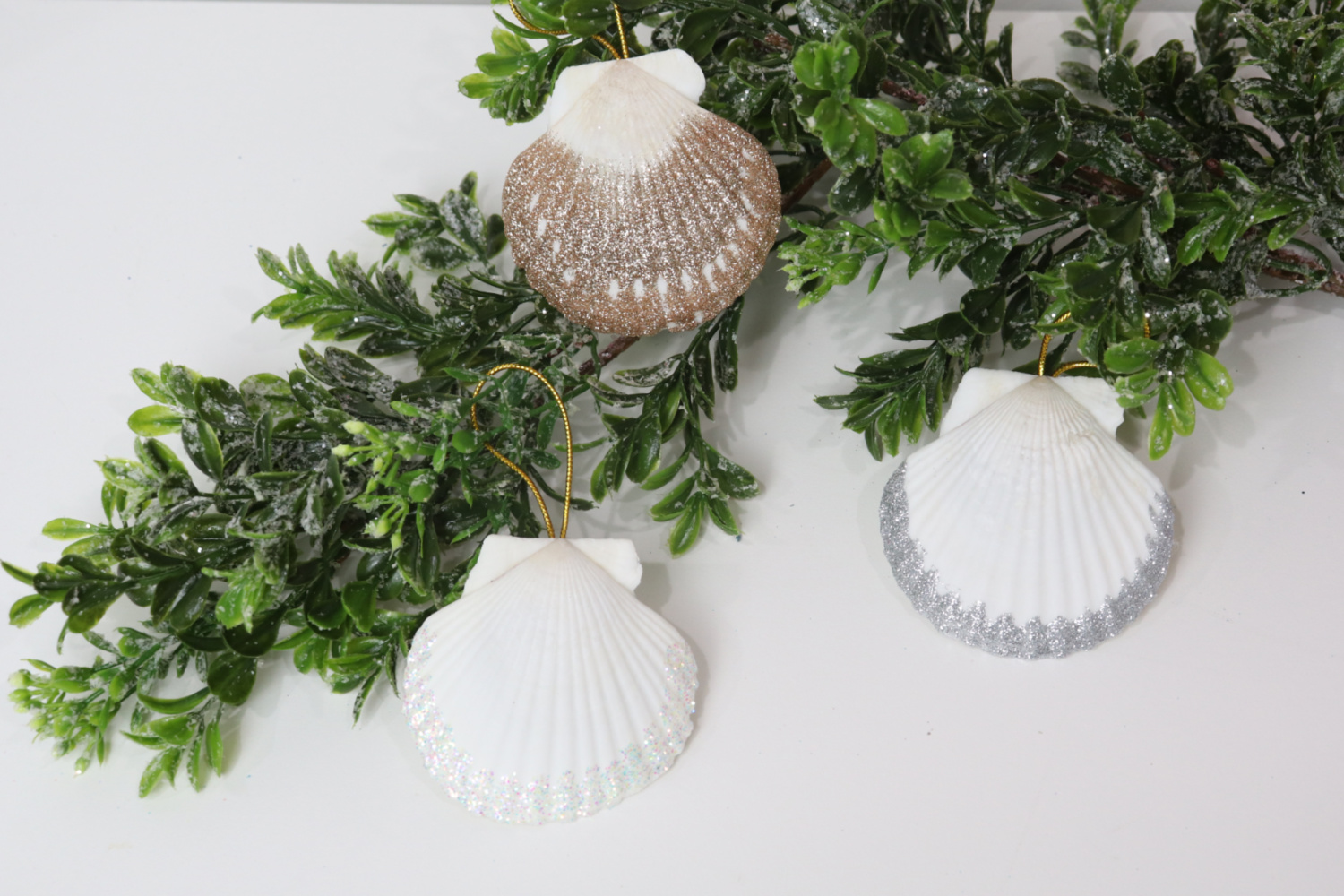 Image includes three glittered seashell ornaments laying on a faux branch on top of a white table.