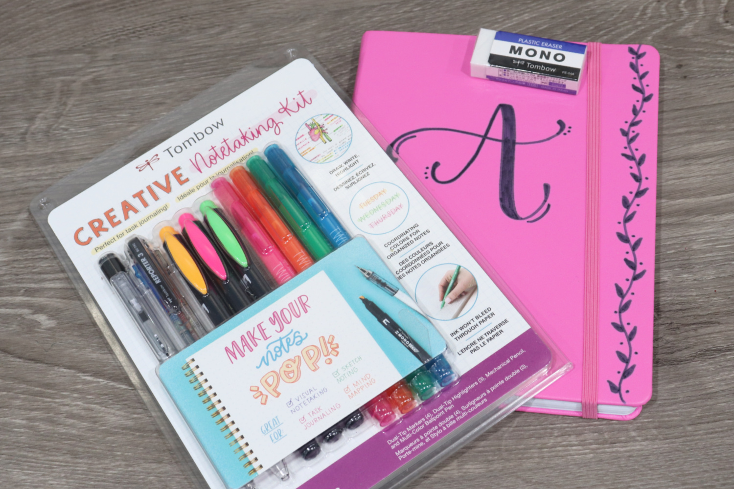 Image contains a pink notebook laying on a wooden desk with a packaged Tombow Creative Notetaking Kit on top. 