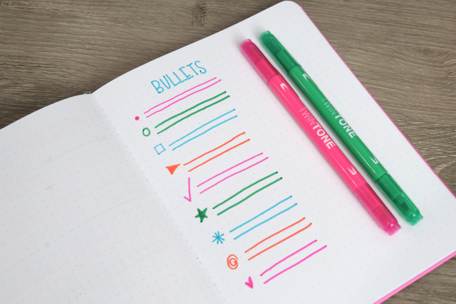 Image contains an open notebook on a wooden desk. On the page are a variety of bullet shapes. A pink and a green marker sit nearby.