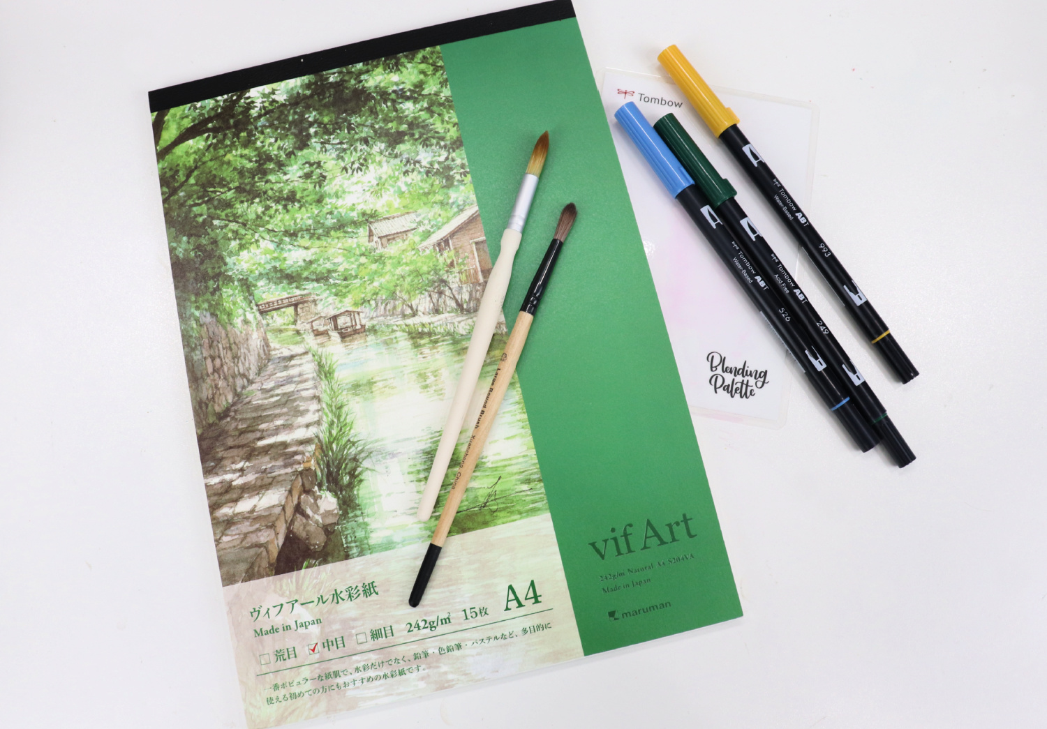 Image contains a watercolor pad with a green cover, a blending palette, three markers, and two paintbrushes on a white background.